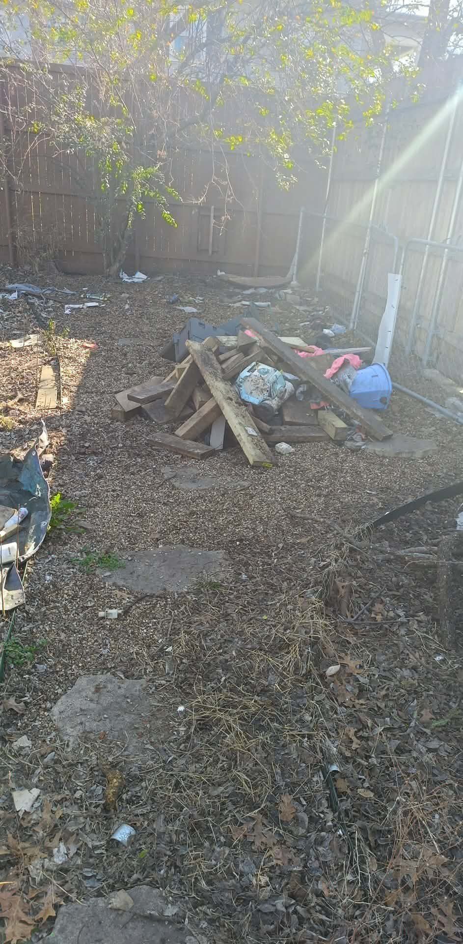 A pile of scrap wood and miscellaneous debris sits in the center of a backyard covered in dead leaves.