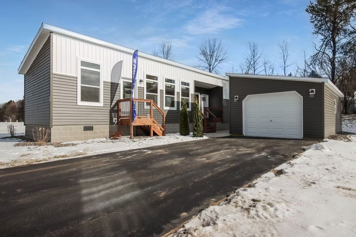 Mobile home with attached garage, asphalt driveway, and snow-covered ground, under a blue sky.