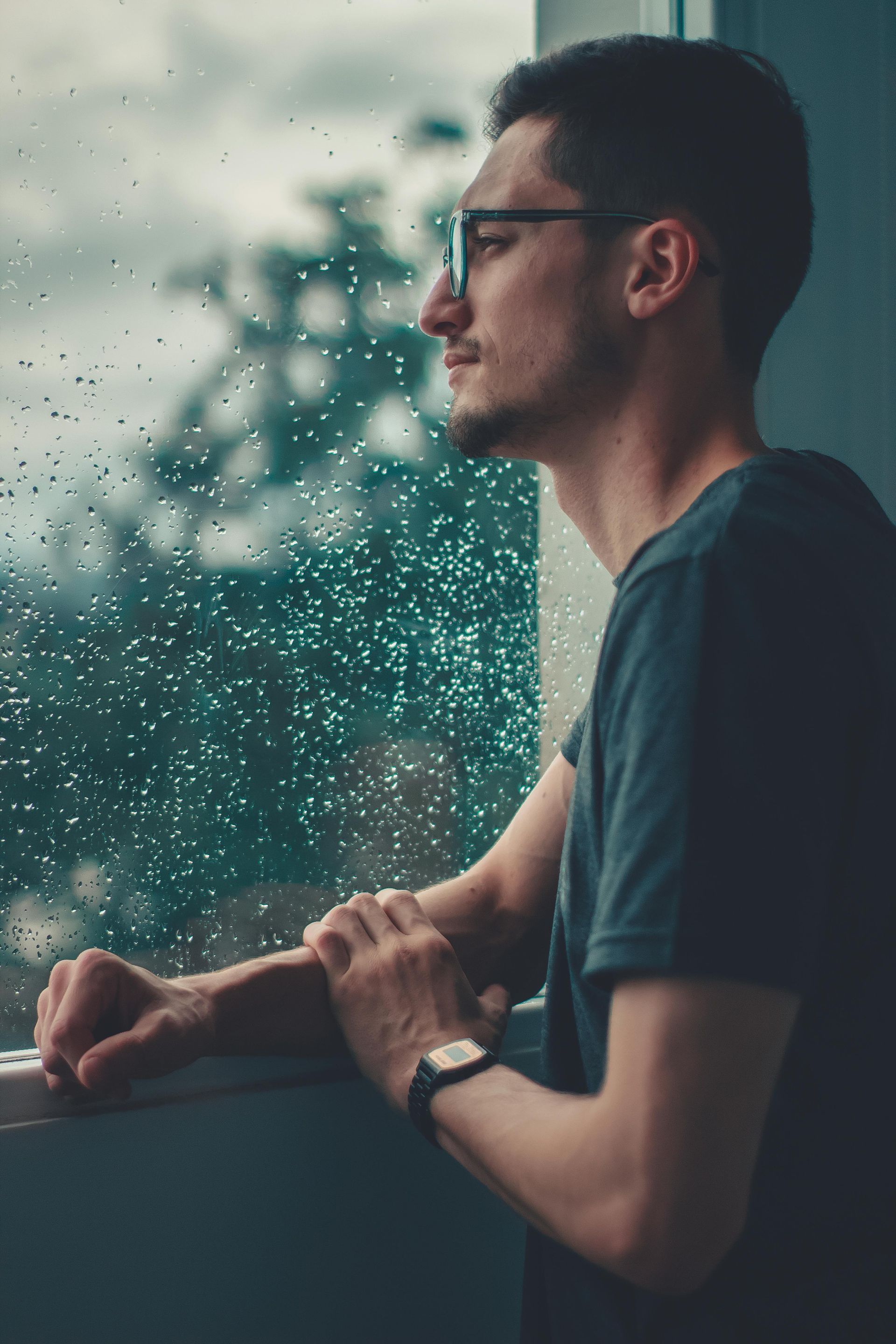 A man wearing glasses is looking out of a window on a rainy day.