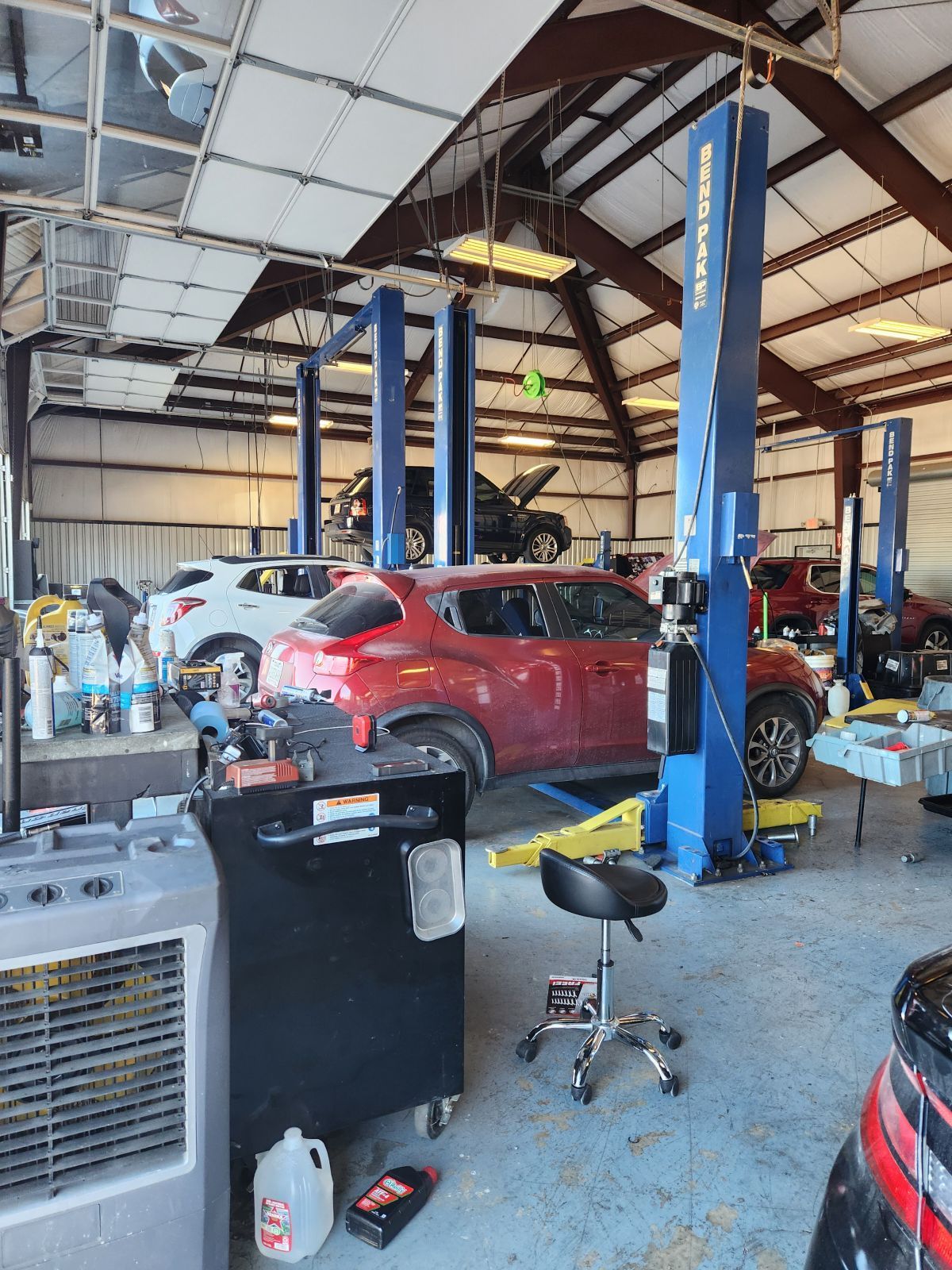 Inside a car repair shop: red SUV on a lift, multiple other vehicles present. Shop has blue lifts and tools. | Mayo Autoworks 2