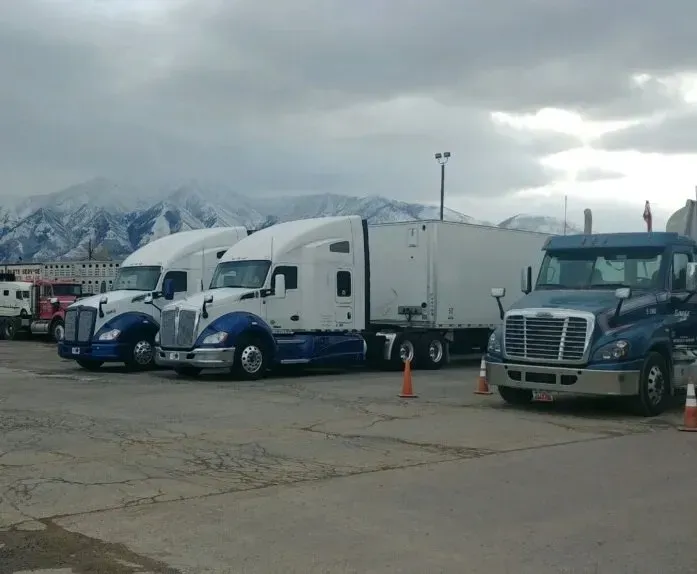 Semi-trucks parked in a lot, with snowy mountains in the background under a cloudy sky.