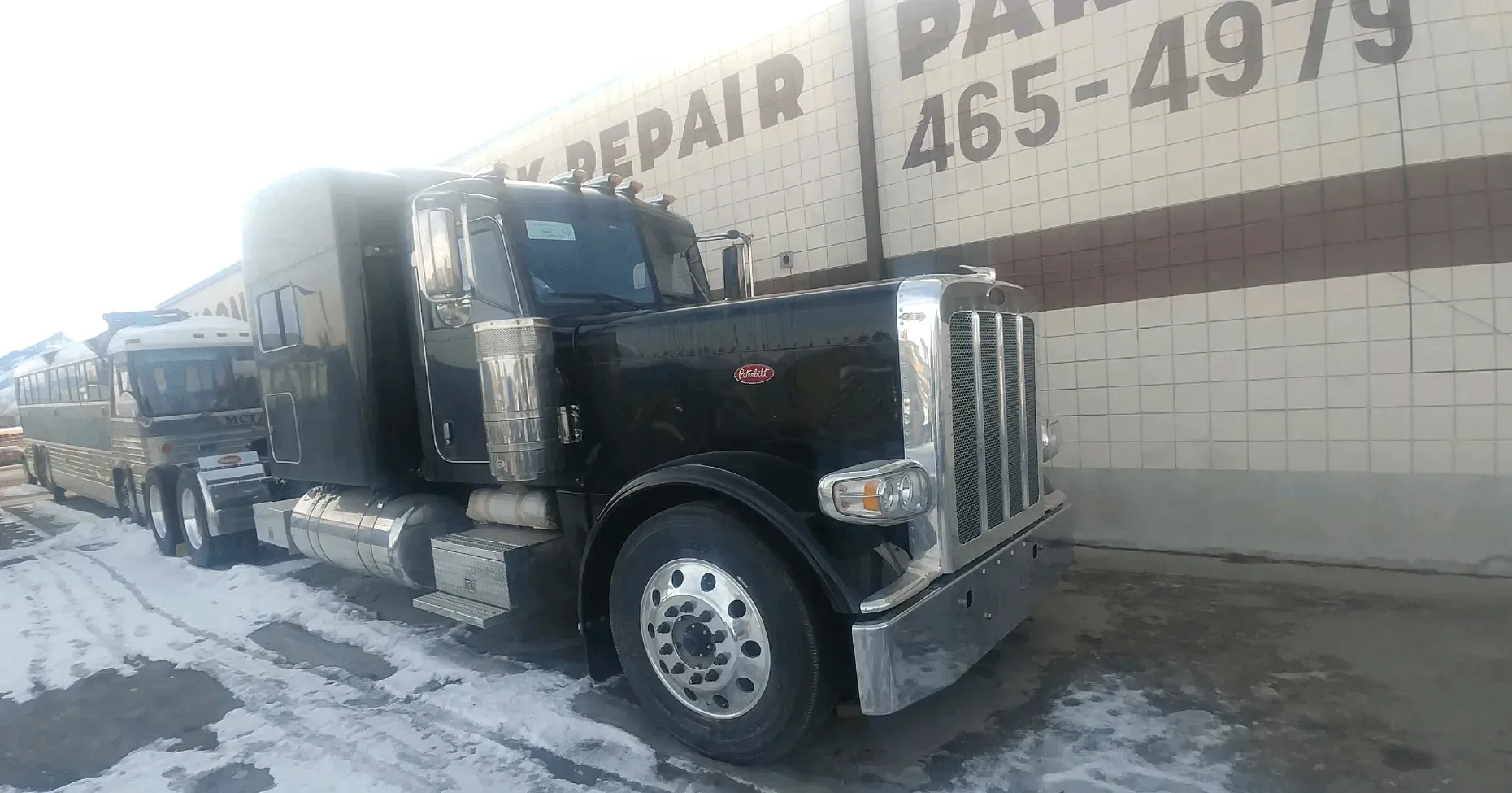 Black semi-truck parked in front of a building with repair signage; snowy ground.