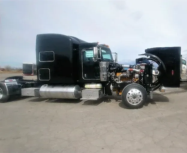 Black semi-truck with the hood open, parked outdoors on a gray surface with blue sky.