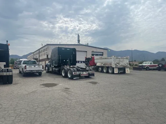 A black semi-truck parked in a lot, beside a white pickup truck and a dump truck, with a building in the background.