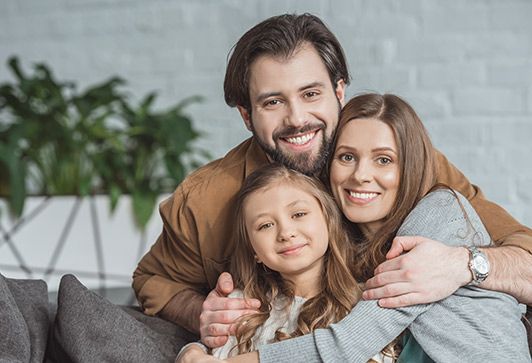 Family of three, a man and woman embracing a young girl, smiling on a gray sofa.