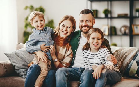 Smiling family of four on a couch: parents, son held in lap, daughter. Warm colors, living room setting.