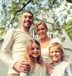 Family of three smiling in front of a brick house; daughter on father's shoulders, mother points.