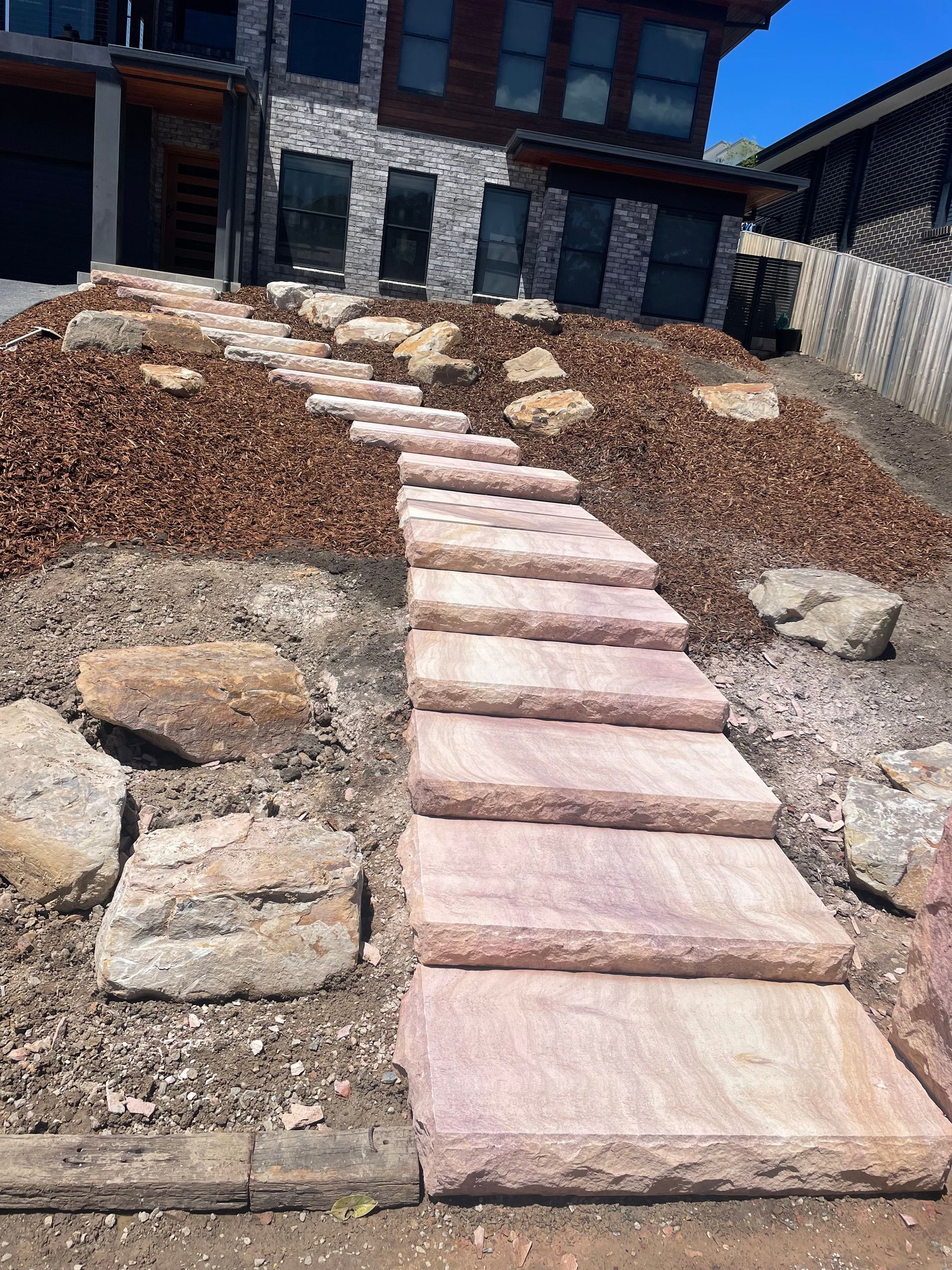 Stone steps lead up a sloped yard to a modern home, surrounded by mulch and rocks.