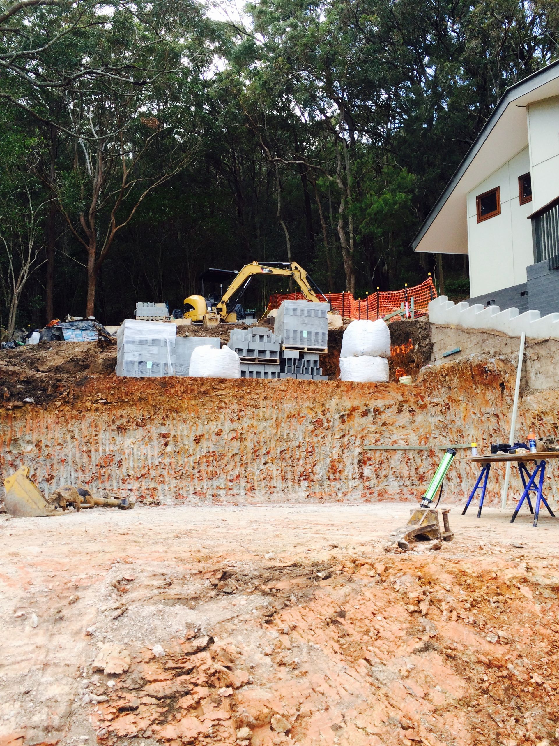 Construction site with excavator, retaining wall, and building. Brown soil and materials against trees.