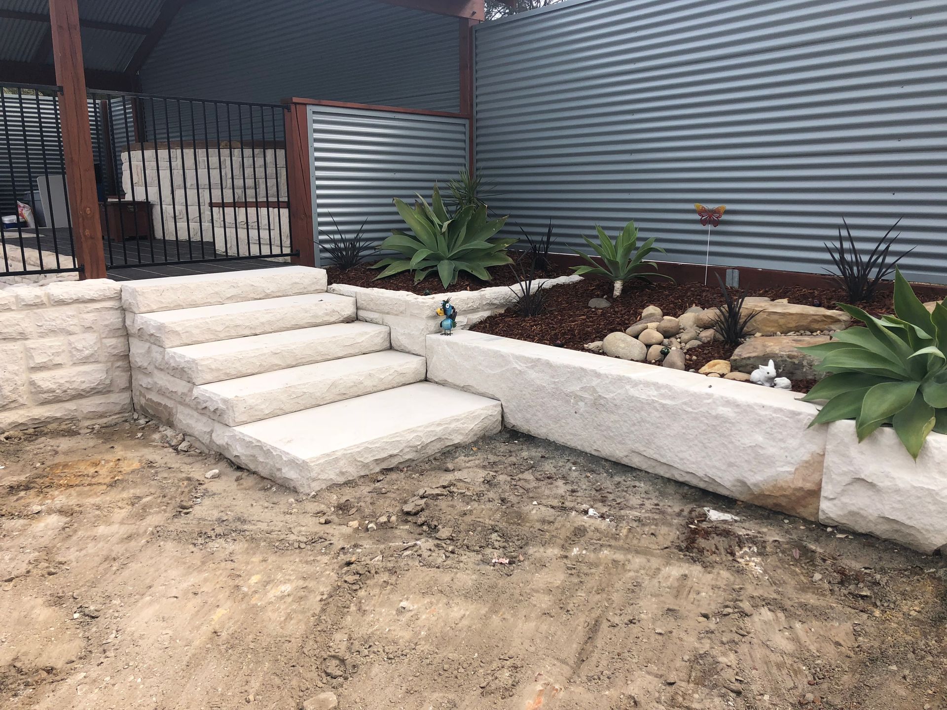 Stone steps lead up to a raised garden bed with plants and corrugated metal wall.