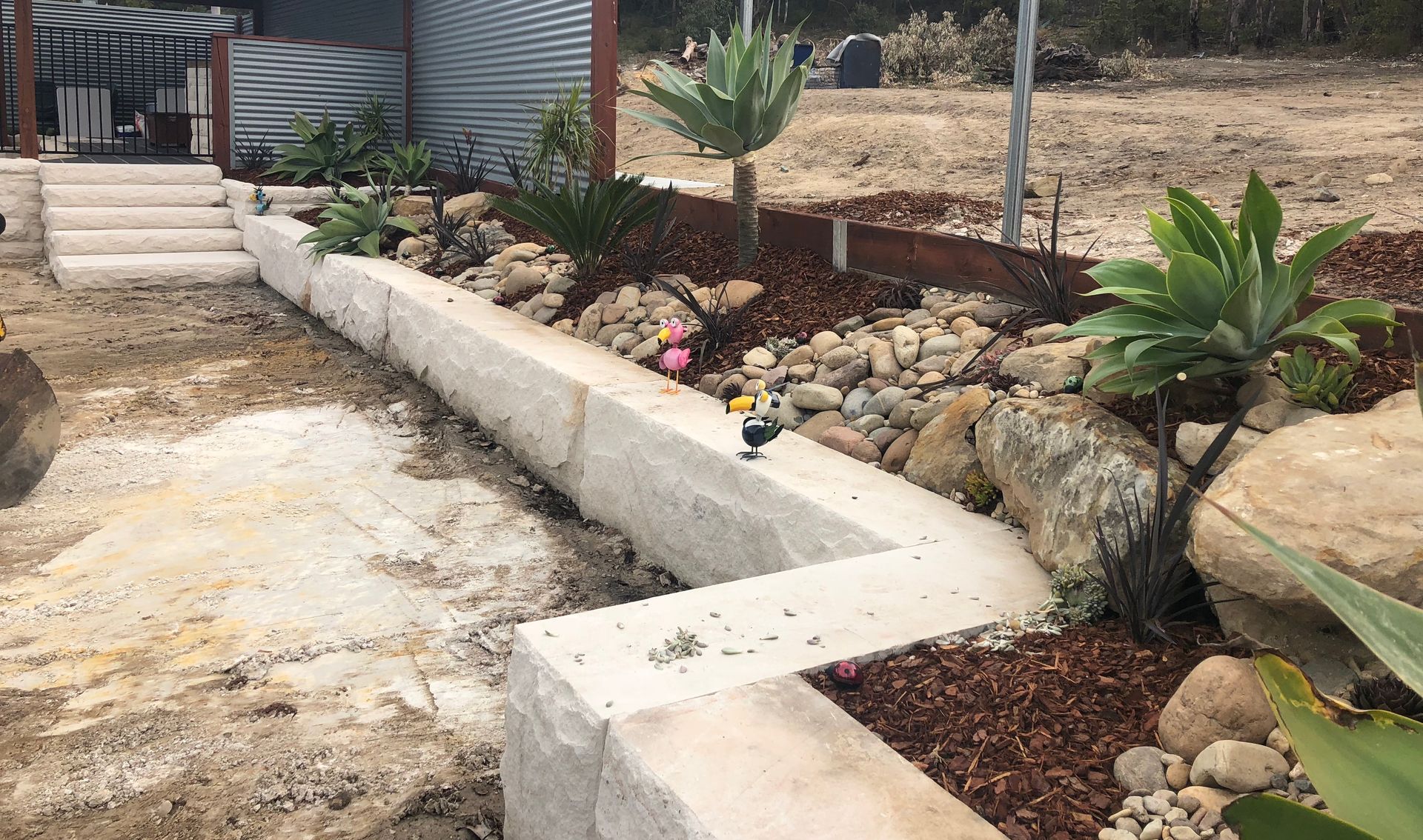Stone retaining wall with landscaped garden, plants, rocks, and wood chips in a sunny outdoor setting.