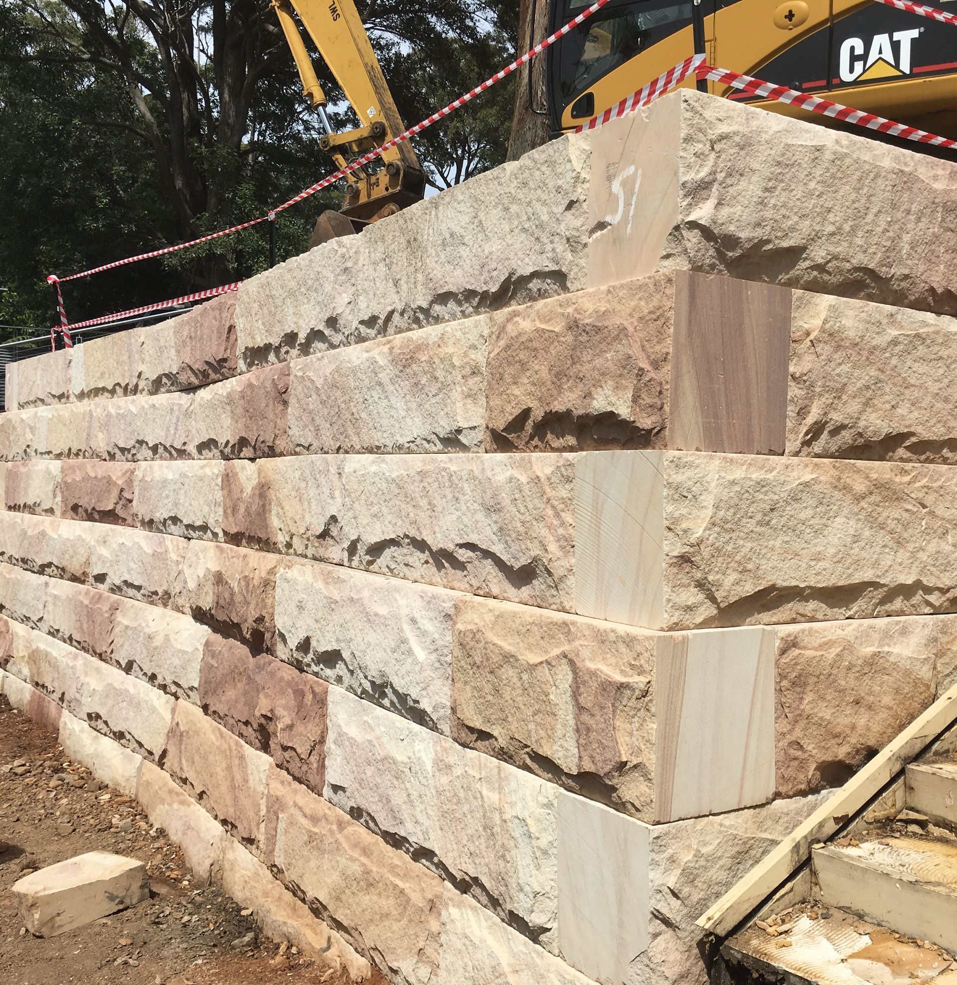 Stone retaining wall being constructed with a yellow excavator in the background.