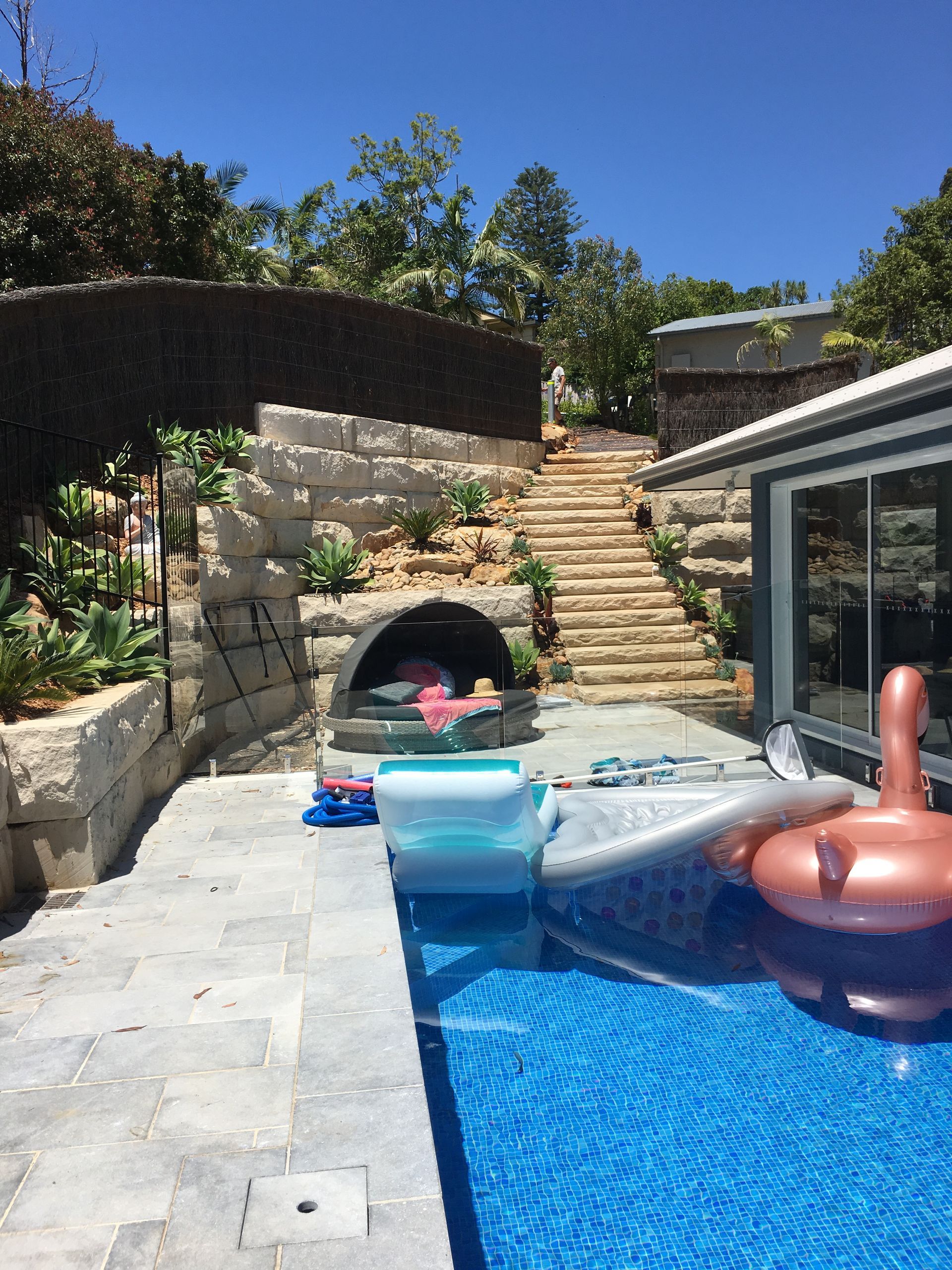 Poolside scene with stone stairs, retaining walls, and a swimming pool. An inflatable flamingo floats in the pool.