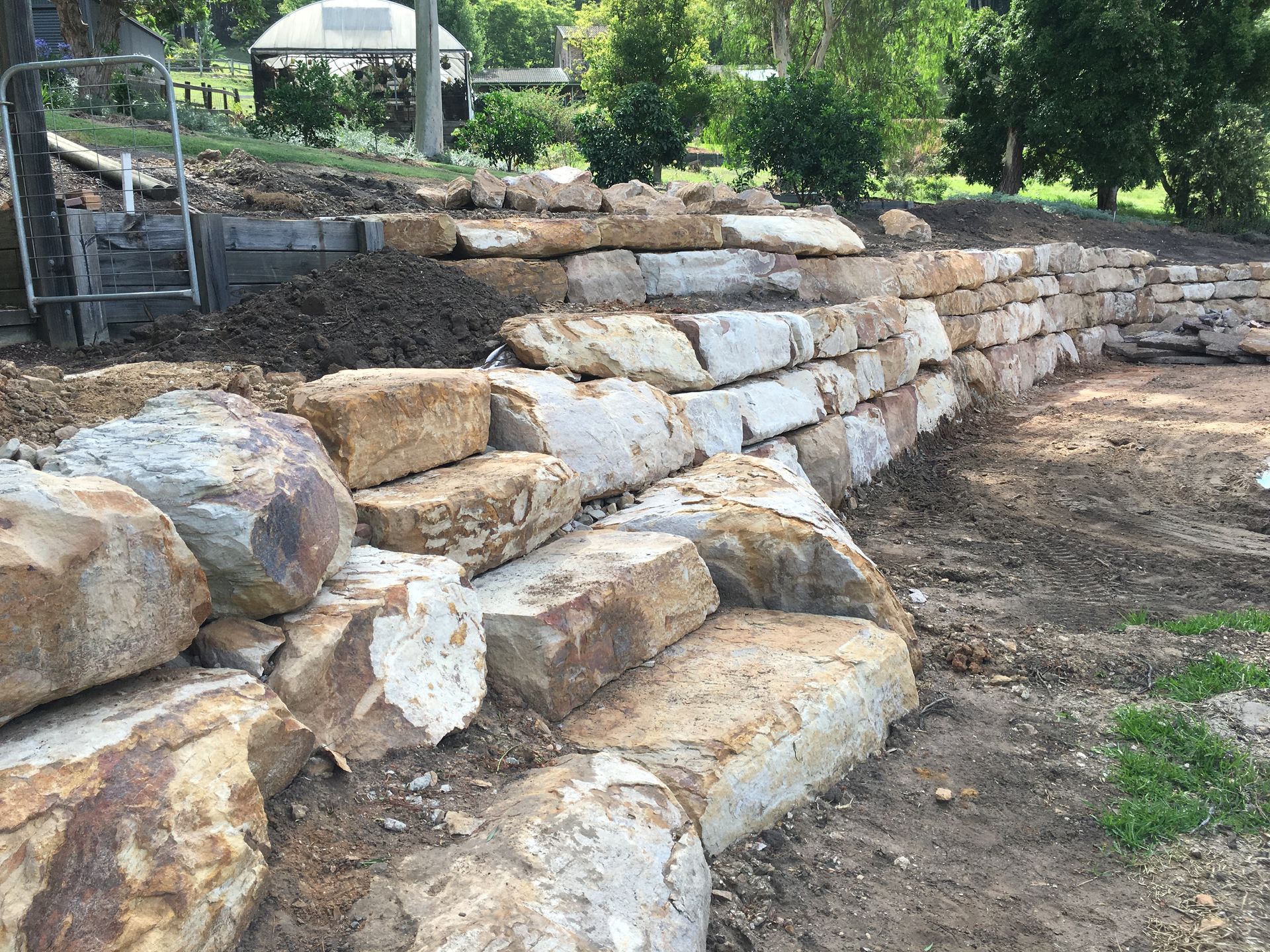 Stone retaining wall under construction, with large, light-colored blocks, soil, and a green grassy area.