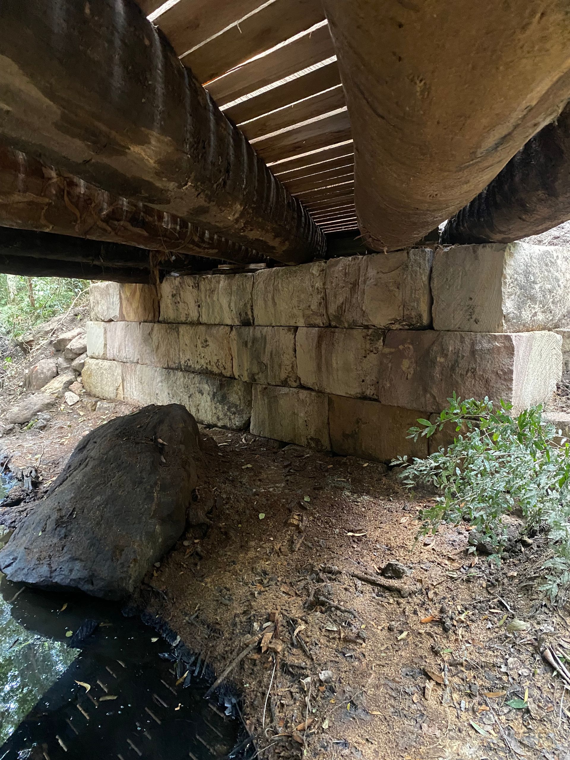 View from beneath a weathered concrete bridge with a brick support over a small stream.