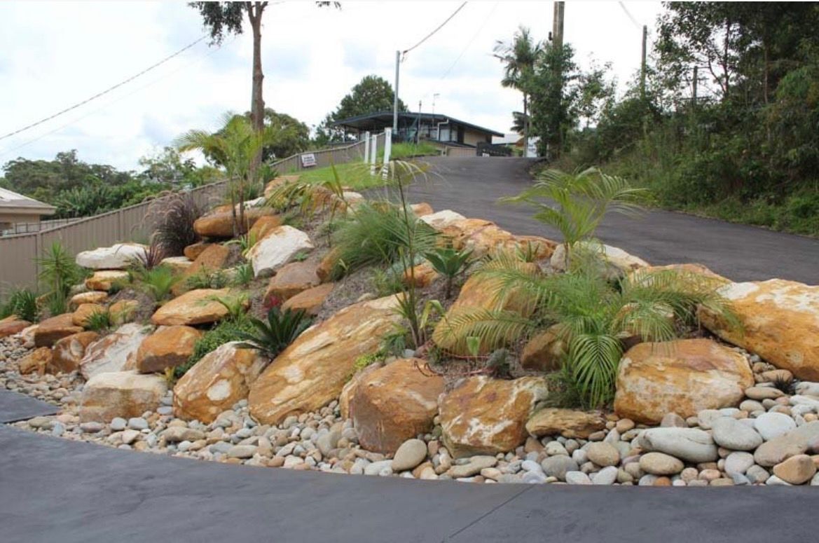 Rocky landscaping with boulders and plants bordering a driveway leading uphill towards a house.