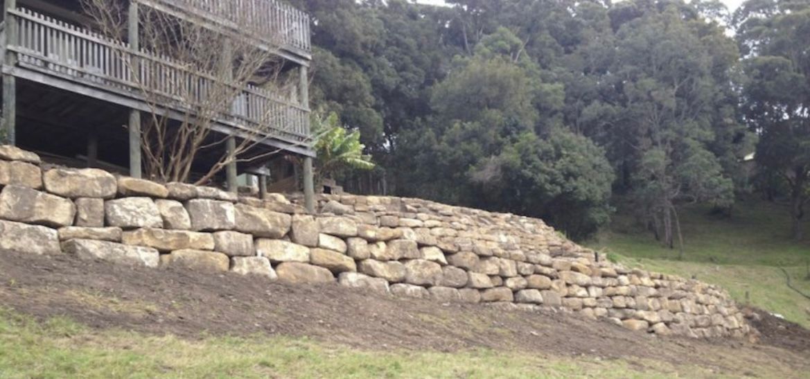 A two-story wooden house on a hillside with a rock retaining wall. Lush green trees fill the background.
