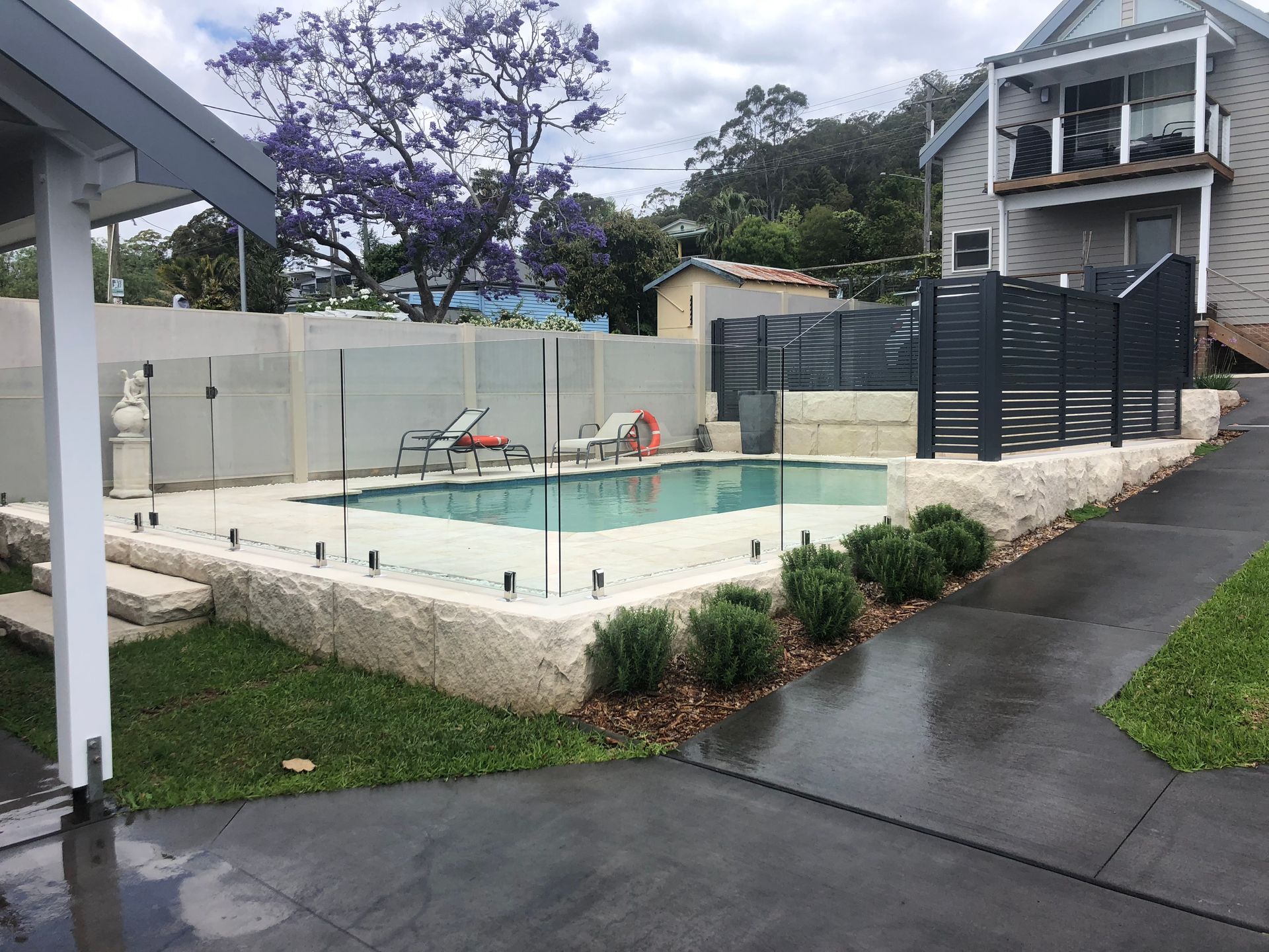 Pool with glass fence surrounded by light-colored stone, bushes, and a two-story house.
