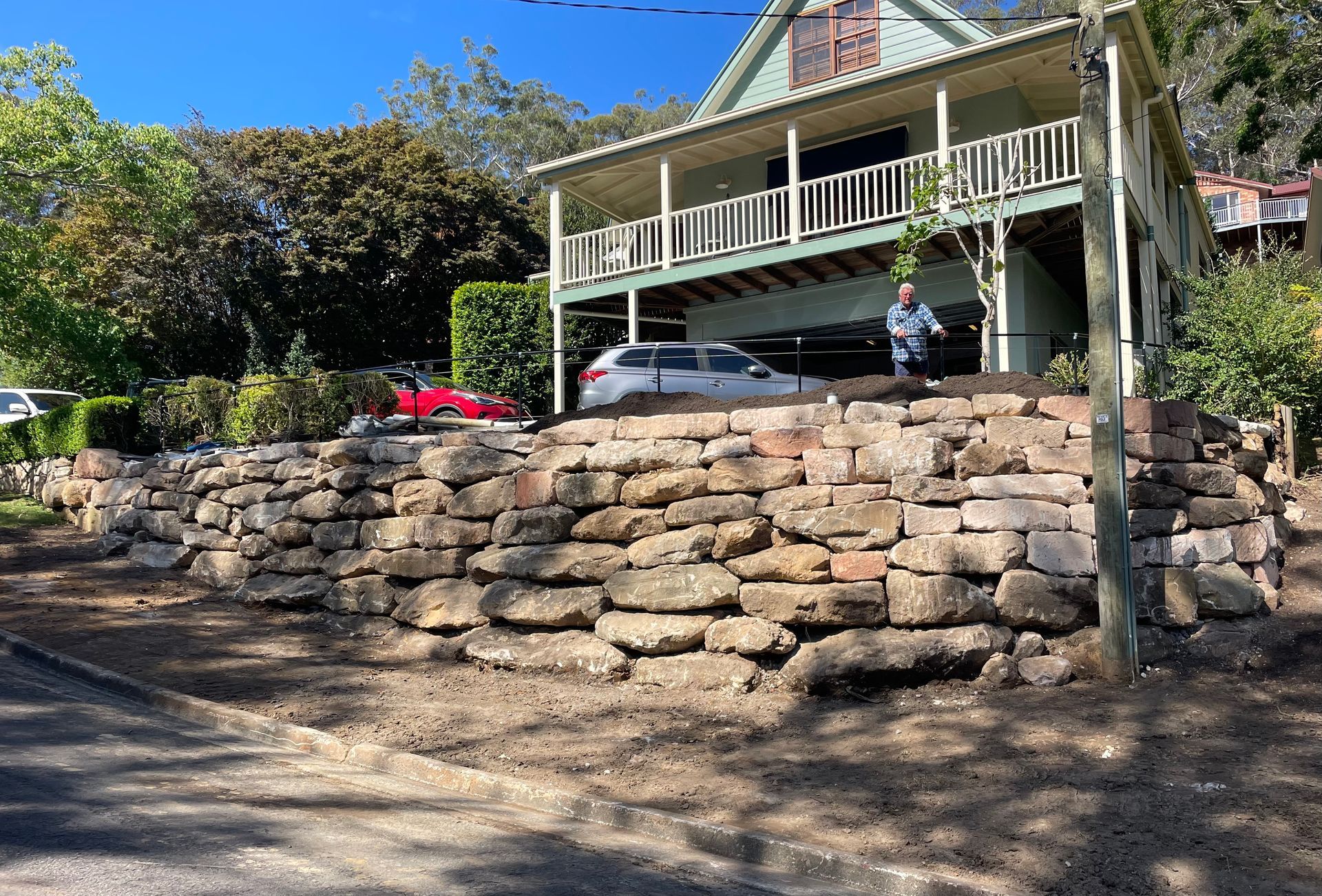 A house on a hill with a stone retaining wall. A car is parked under the balcony. Person standing on the porch.