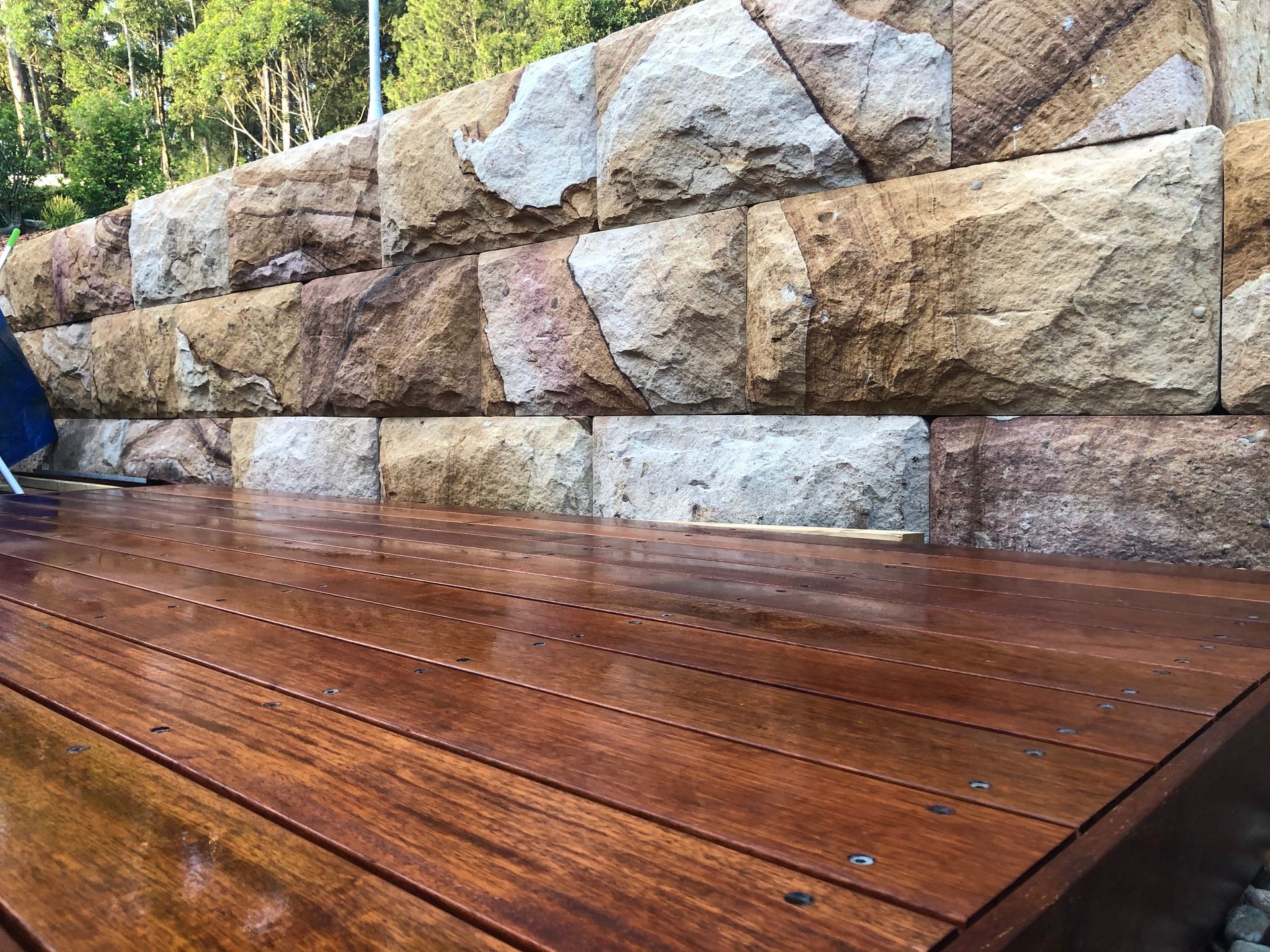 Wet, dark-stained wooden deck in foreground, sandstone retaining wall in background; outdoor setting.