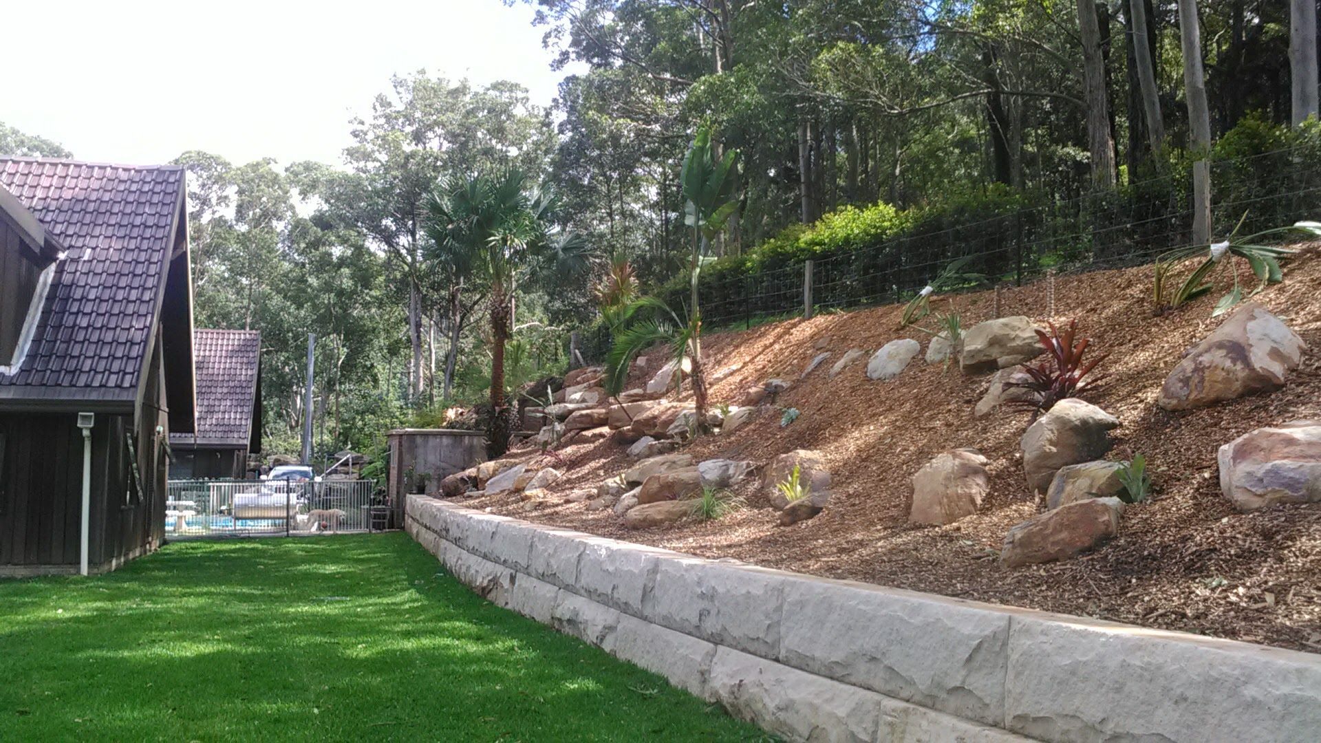 Stone retaining wall borders grassy lawn and leads up to a sloped, rocky hillside with trees.