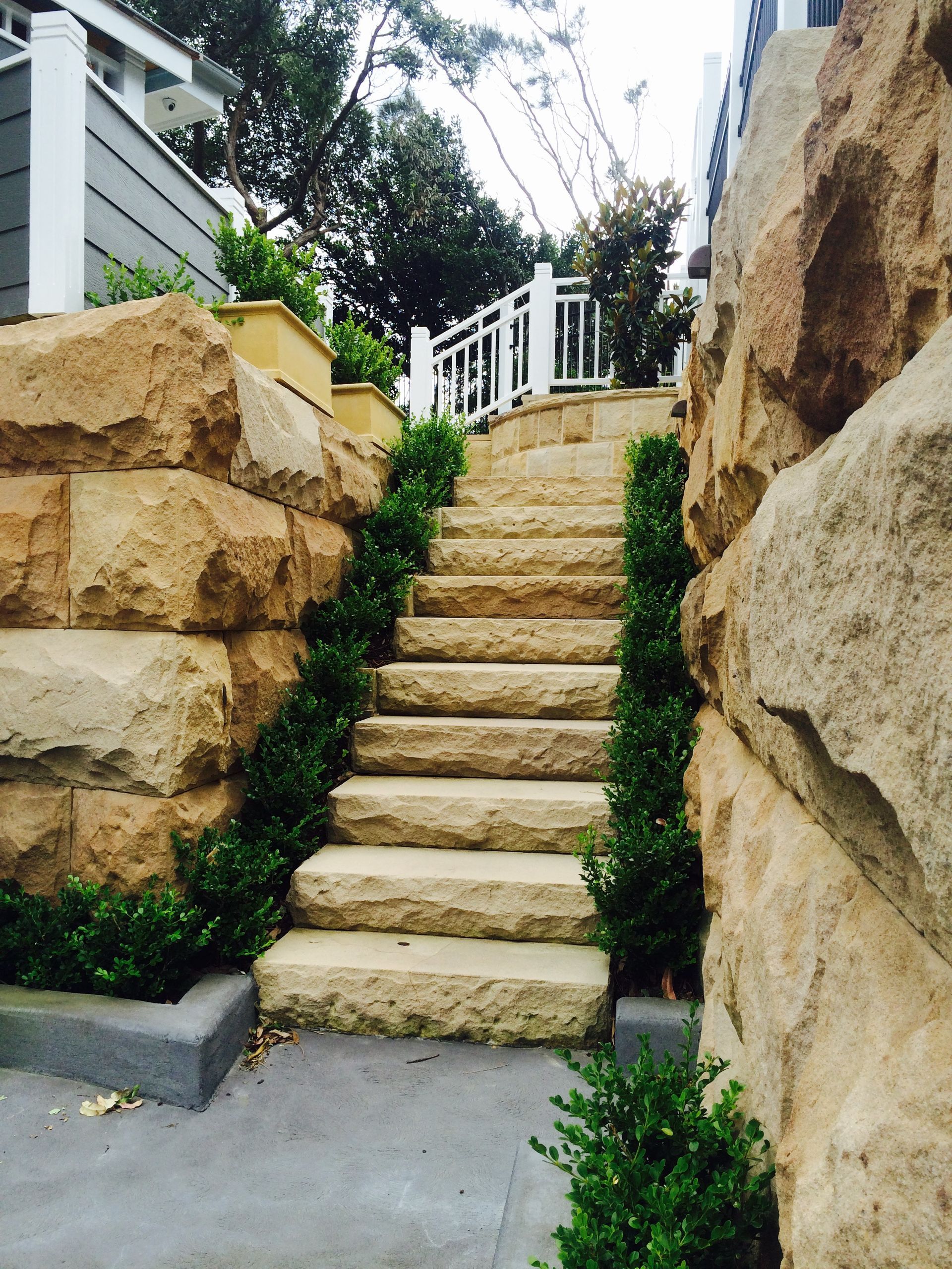 Stone steps flanked by rock walls and green bushes lead to a white railing.