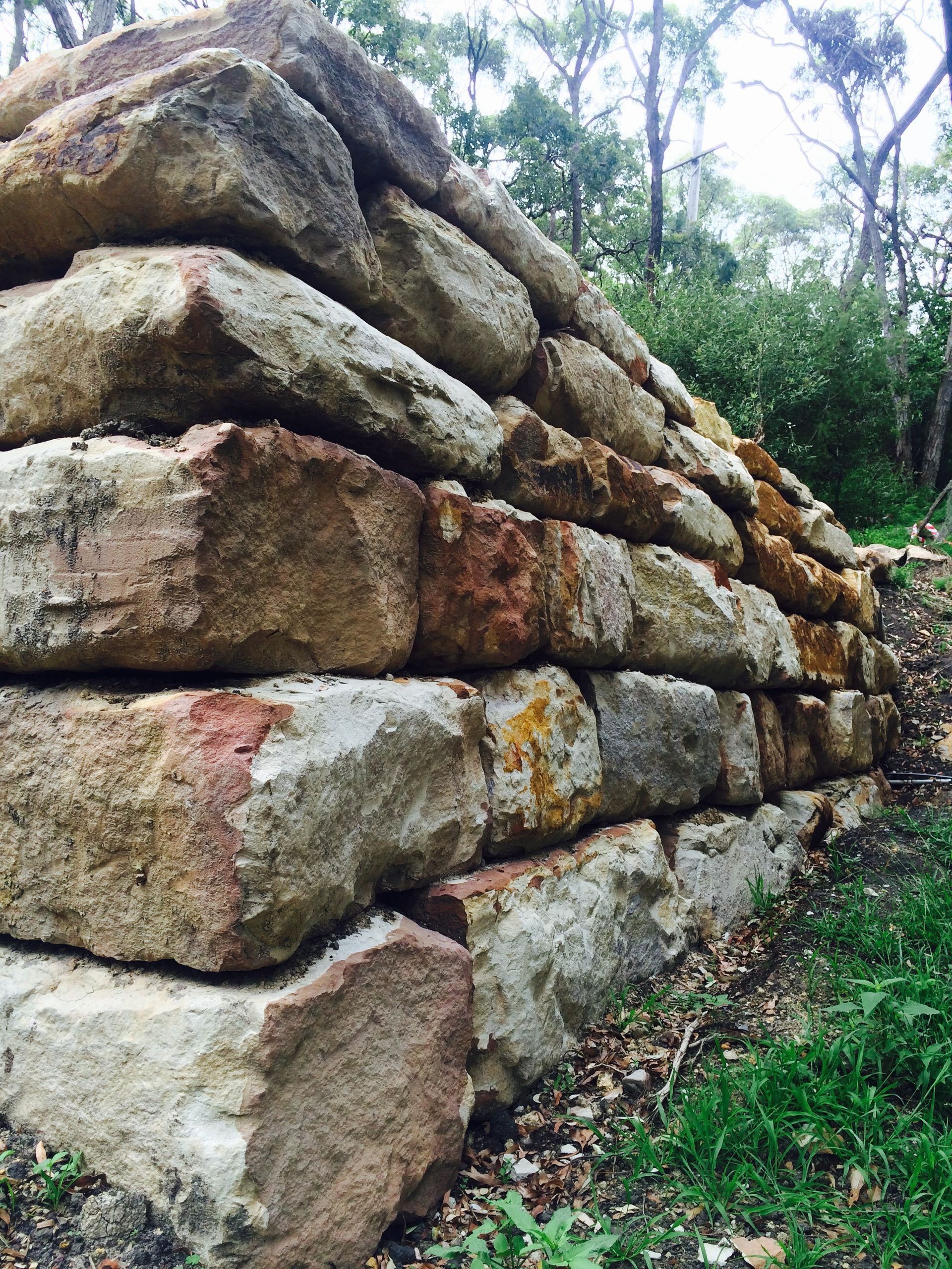 Stone retaining wall, made of stacked rectangular blocks, in a wooded area with grass.
