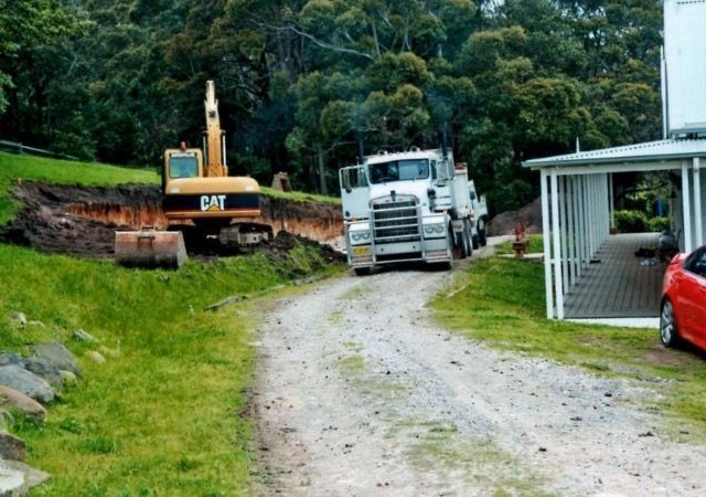 Excavator and truck on a gravel driveway next to a building and a grassy hill.