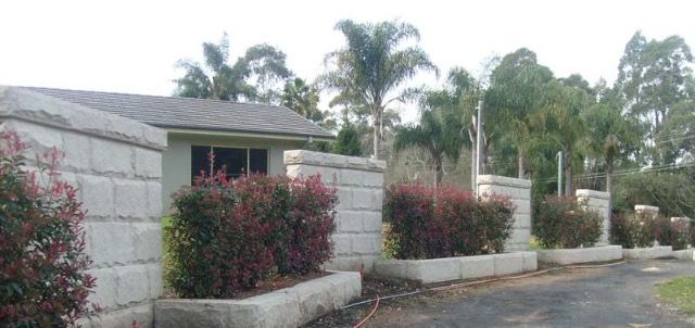 Gray concrete wall with red bushes, small building, trees.