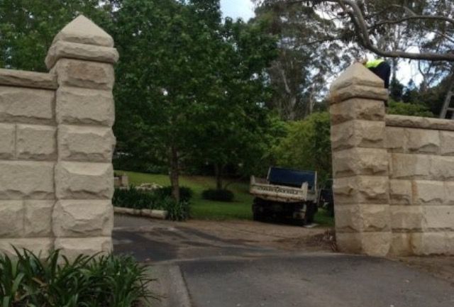 Stone entrance pillars frame a driveway; a truck sits inside, a person on a ladder to the right.
