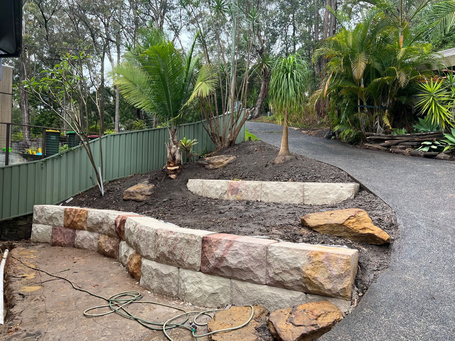 Stone retaining wall with a mulch-covered garden bed, a gravel driveway, and trees.