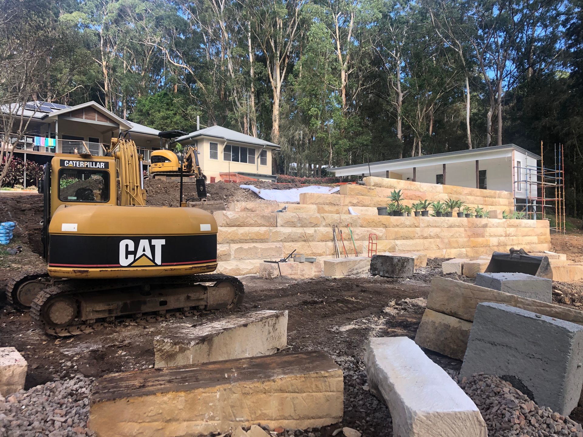 Yellow CAT excavator on construction site with retaining wall and buildings in background.