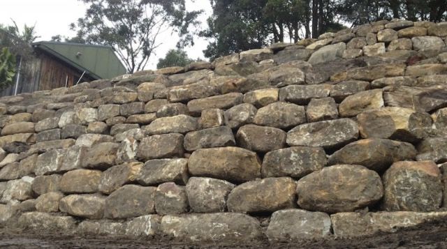 Stone retaining wall built on a hillside. Houses and trees visible in background.