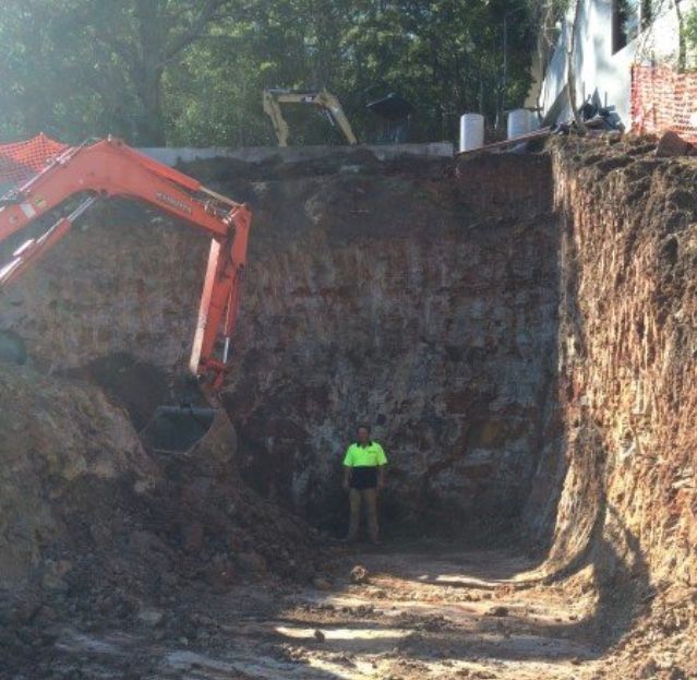 Construction worker standing in a deep excavated trench; orange excavator beside him.