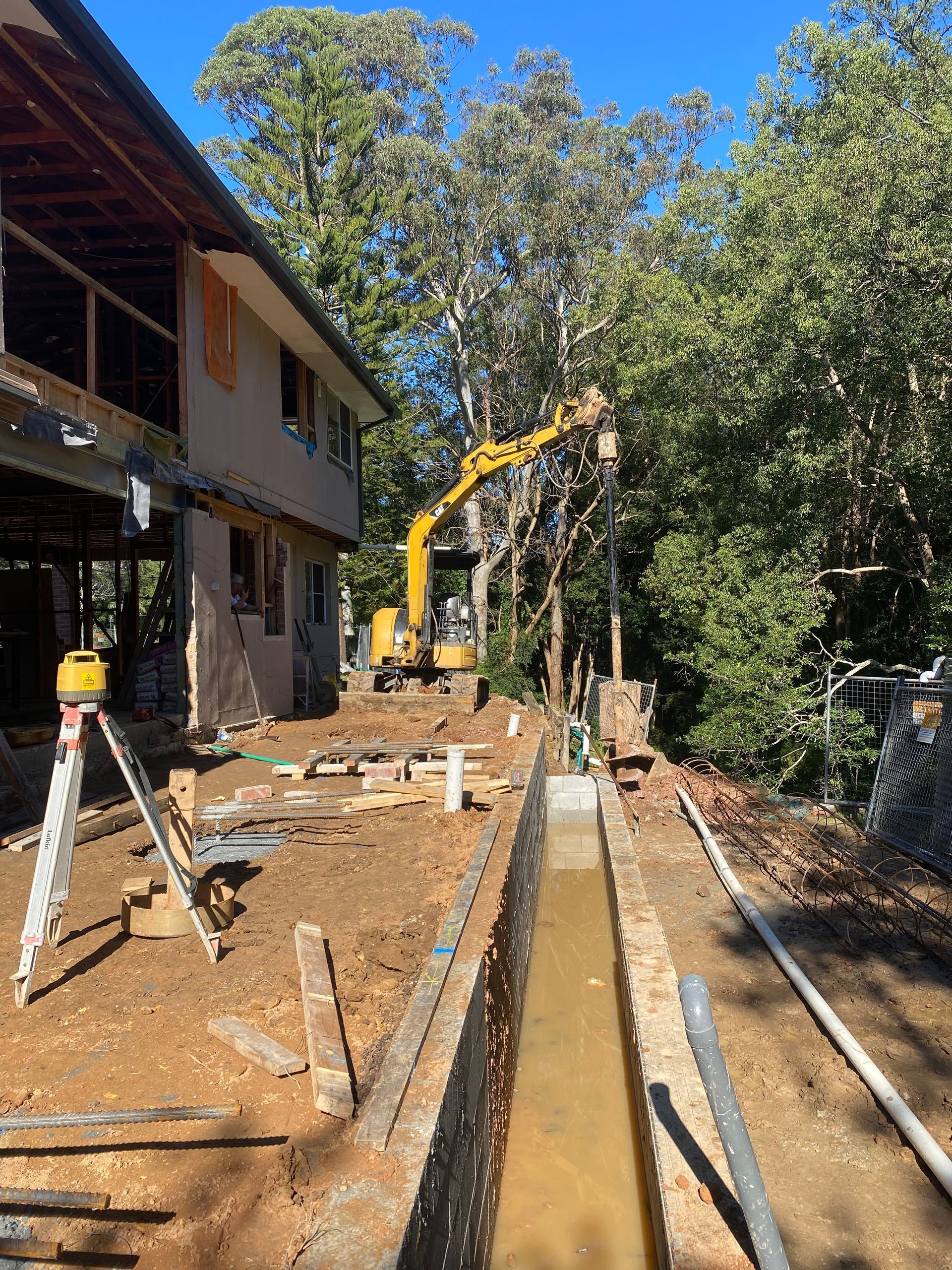 Construction site: excavator operating near a building and trench. Dirt and construction materials are visible.
