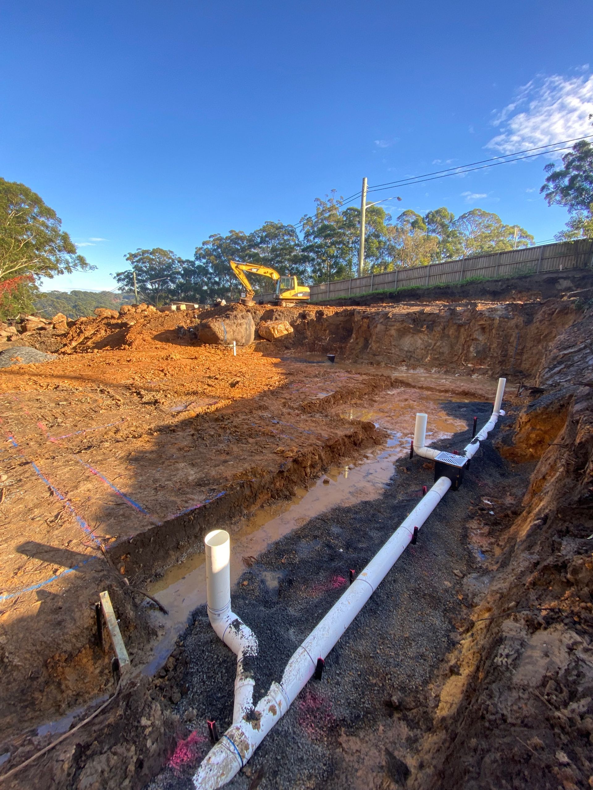 Construction site with white PVC pipes in a muddy trench; excavator in background.