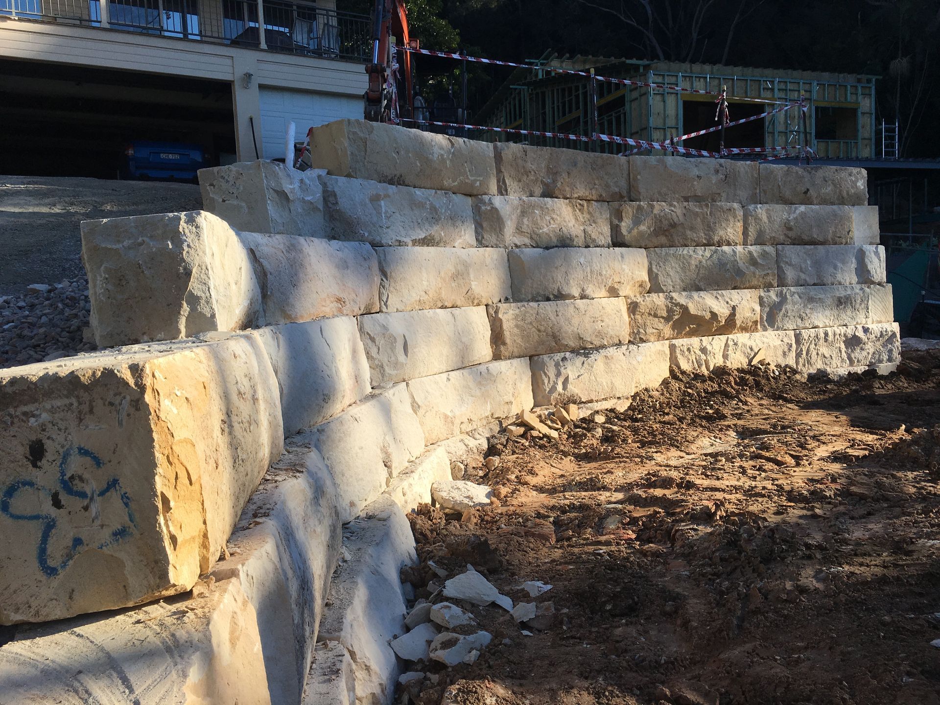 Retaining wall built of light-colored stone blocks, on a hillside, with a building under construction in the background.