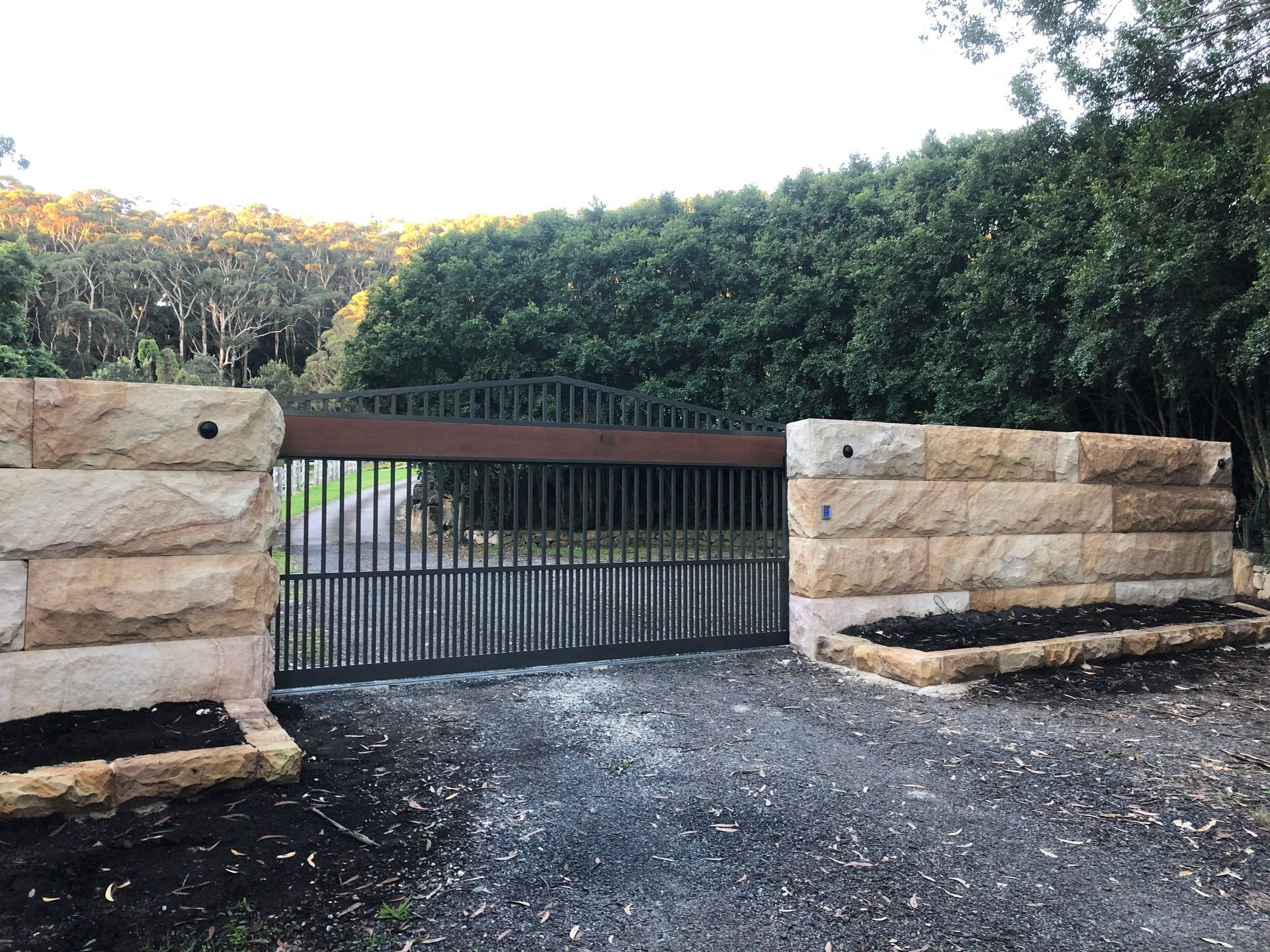 Stone gate pillars frame a dark metal gate. Gravel driveway and dense green trees form the backdrop.