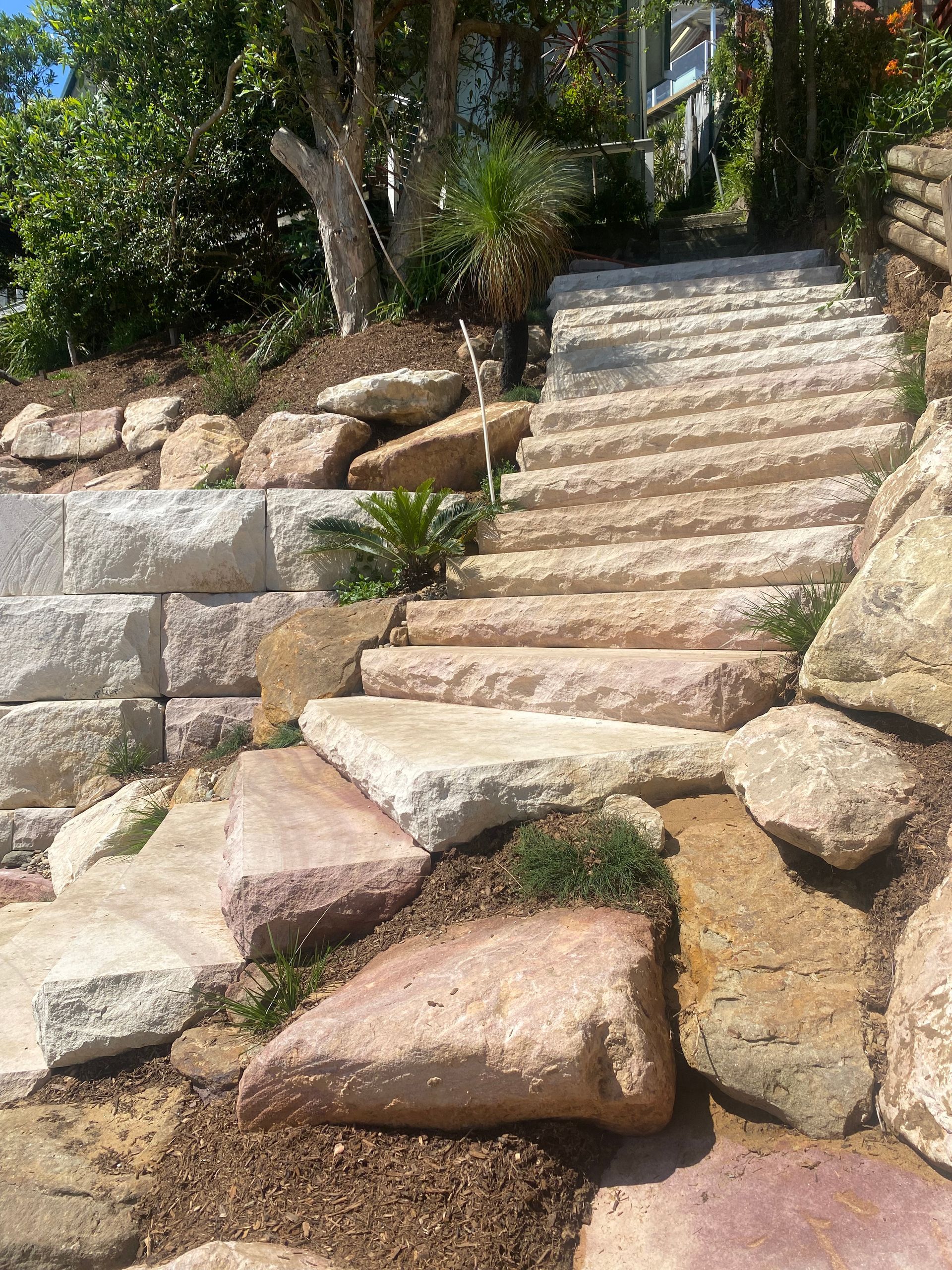 Stone steps ascend a hillside, flanked by large rocks and retaining walls. Sunny outdoor setting.
