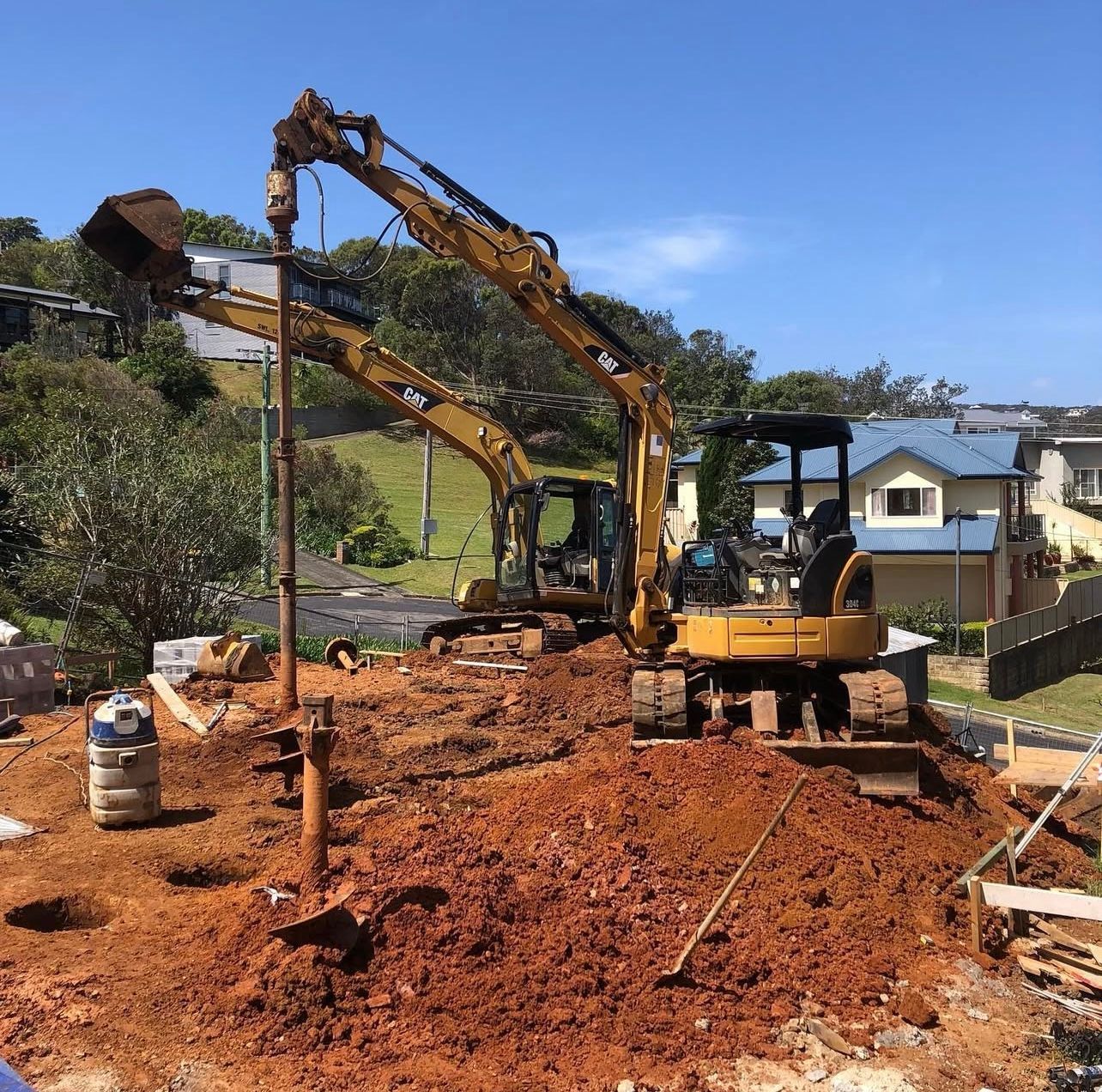 Two yellow excavators drilling holes in red dirt at a construction site near houses.