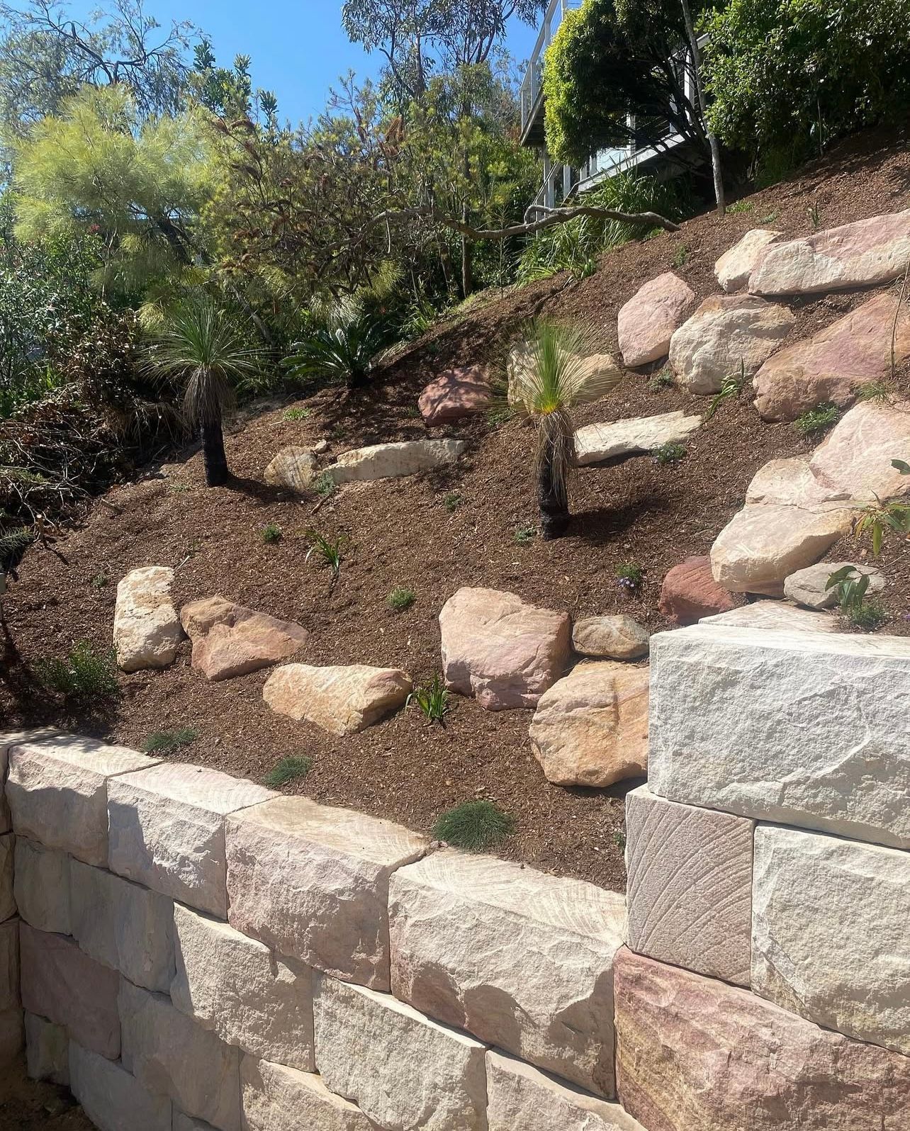 Retaining wall and rockery garden on a hillside, with brown mulch, trees, and light-colored boulders.