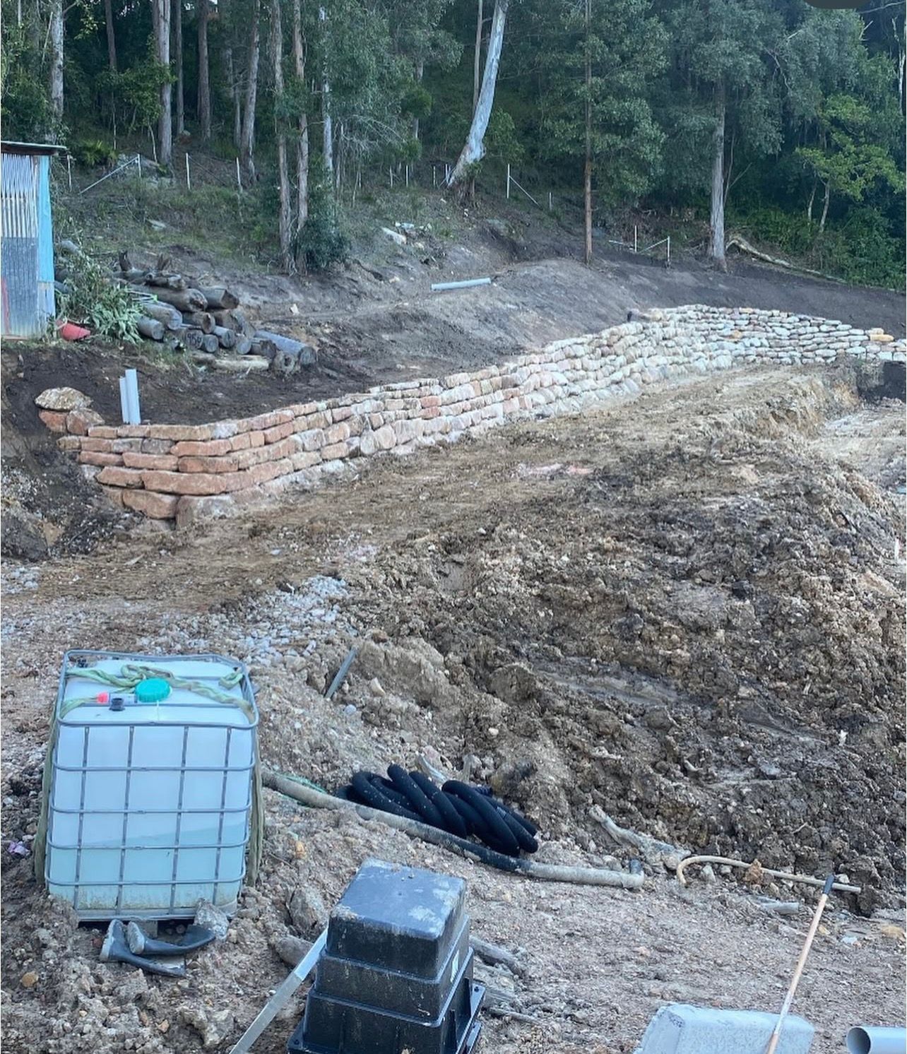 Construction site with wood chip pile, retaining wall, water tank, and trees.