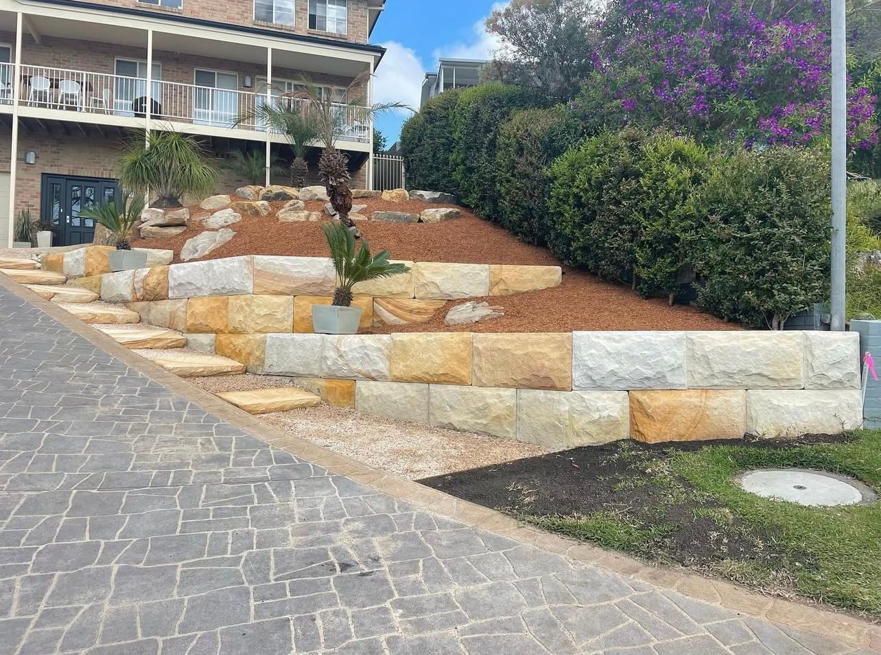Stone retaining walls with stairs, leading up to a landscaped garden bed. A building is in the background.