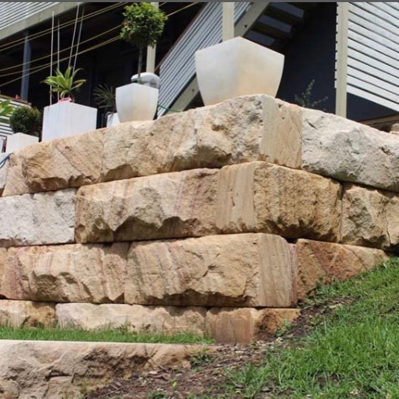 Sandstone retaining wall with potted plants, green grass, and a building in the background.