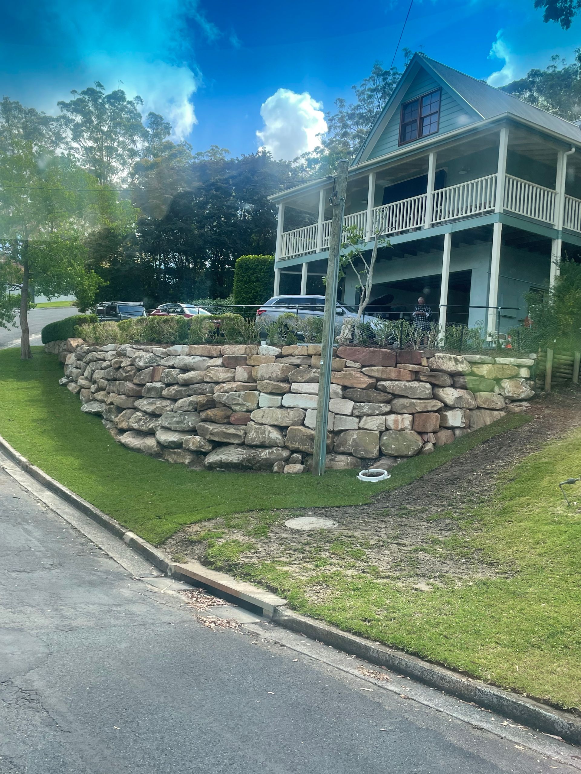 Two-story house with a stone retaining wall along a street on a sunny day.