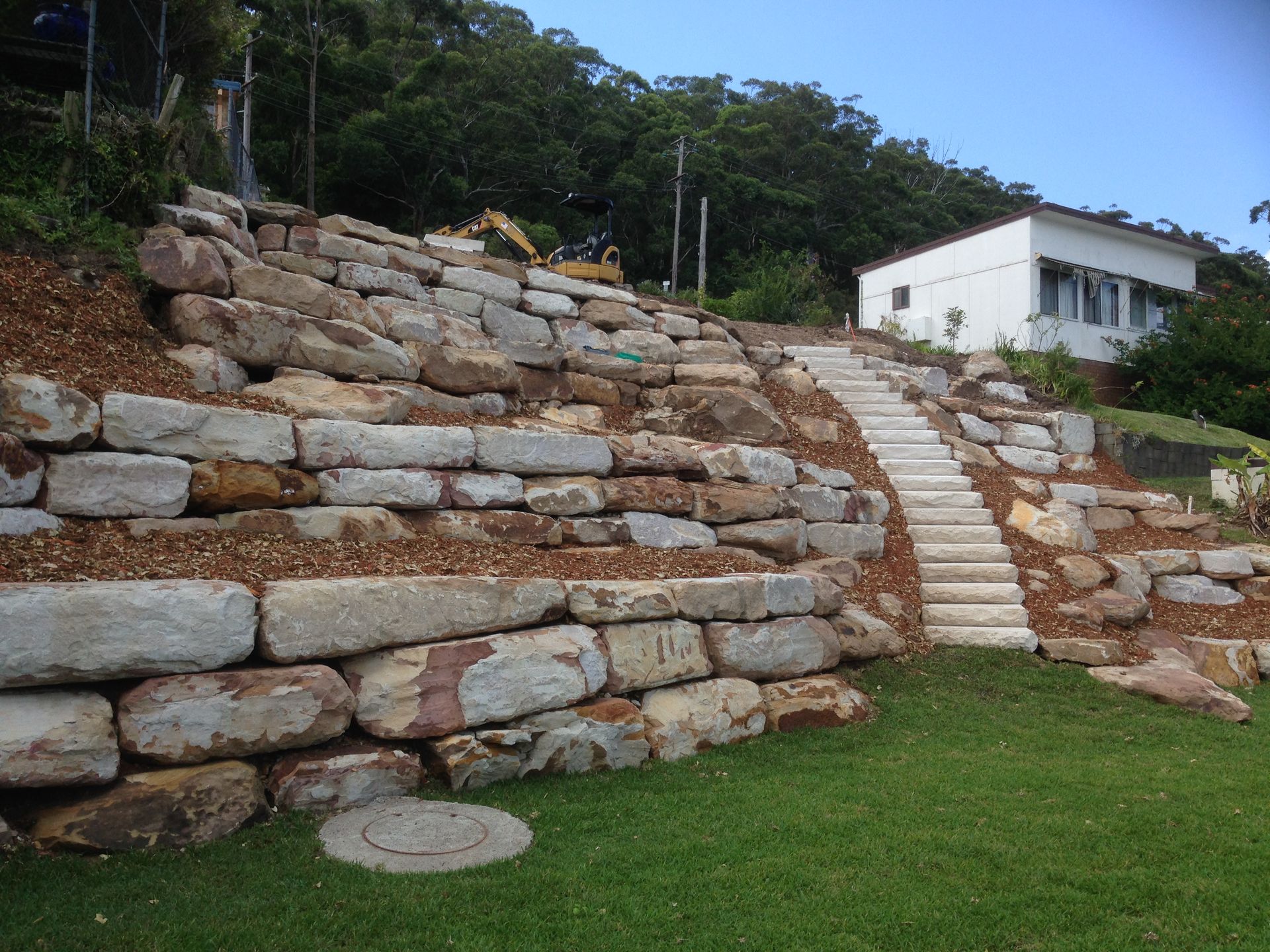 Stone retaining walls with stairs leading to a white house on a hill. Green grass and trees are visible.