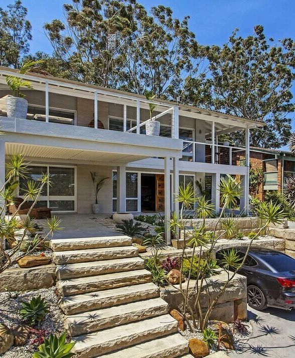 Two-story home with stone steps, carport, and lush landscaping. A car is parked in the driveway.