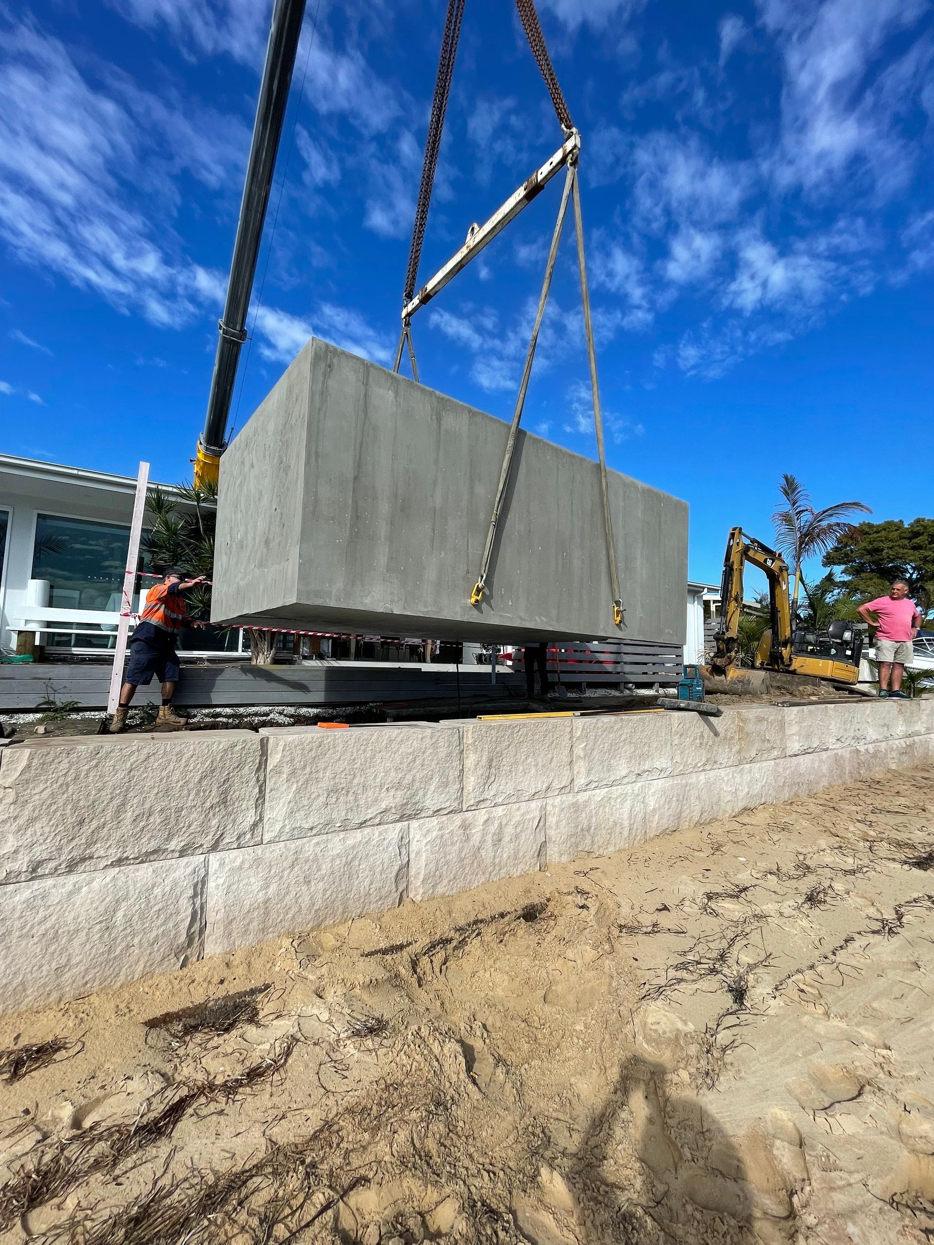 Crane lifting a large concrete block on a beach, construction workers in view.