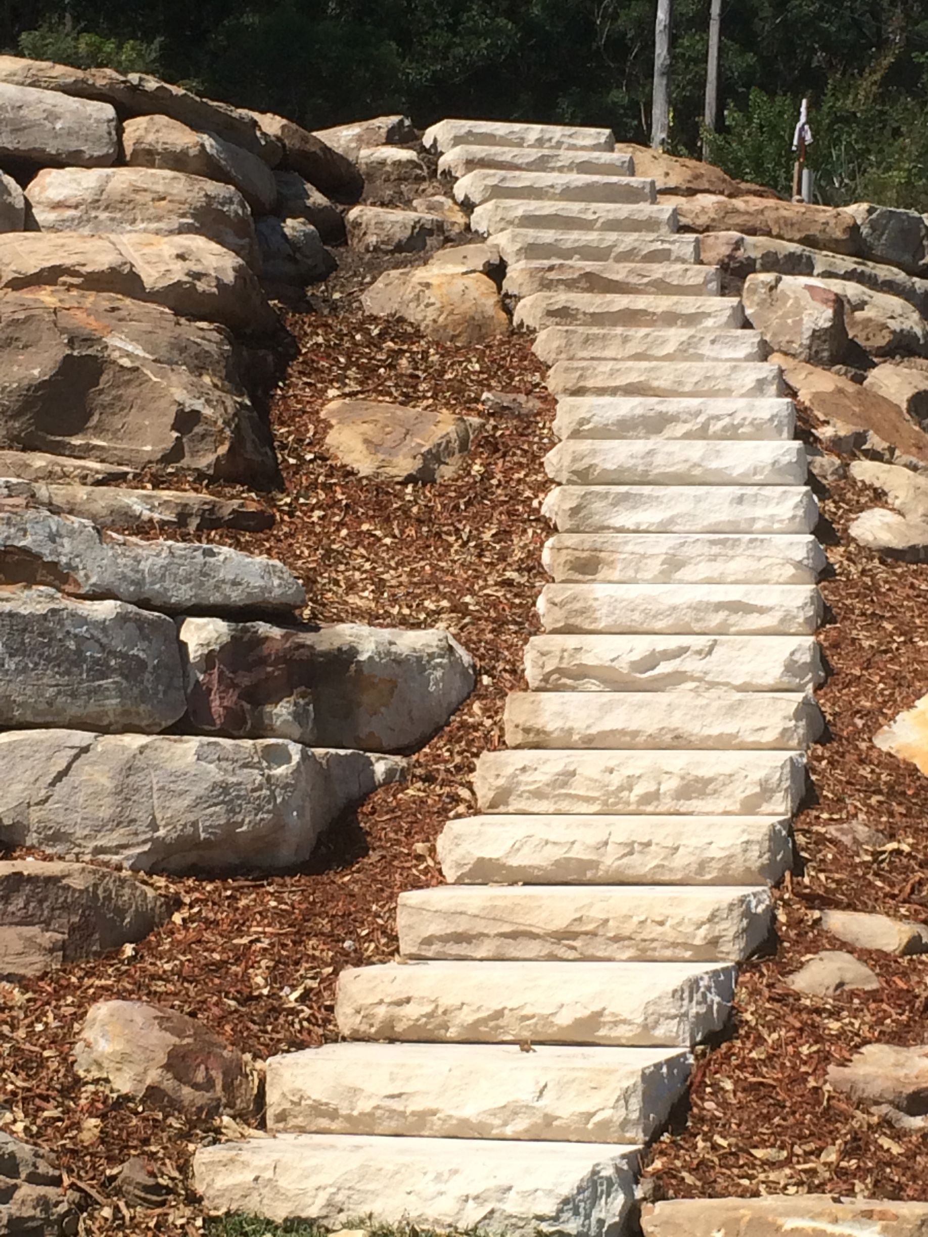 Stone steps ascend a hillside, flanked by rock outcroppings and mulch, leading into a shaded area.