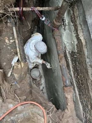 Worker in white protective suit in a narrow trench, examining a pipe beside exposed roots and concrete wall.